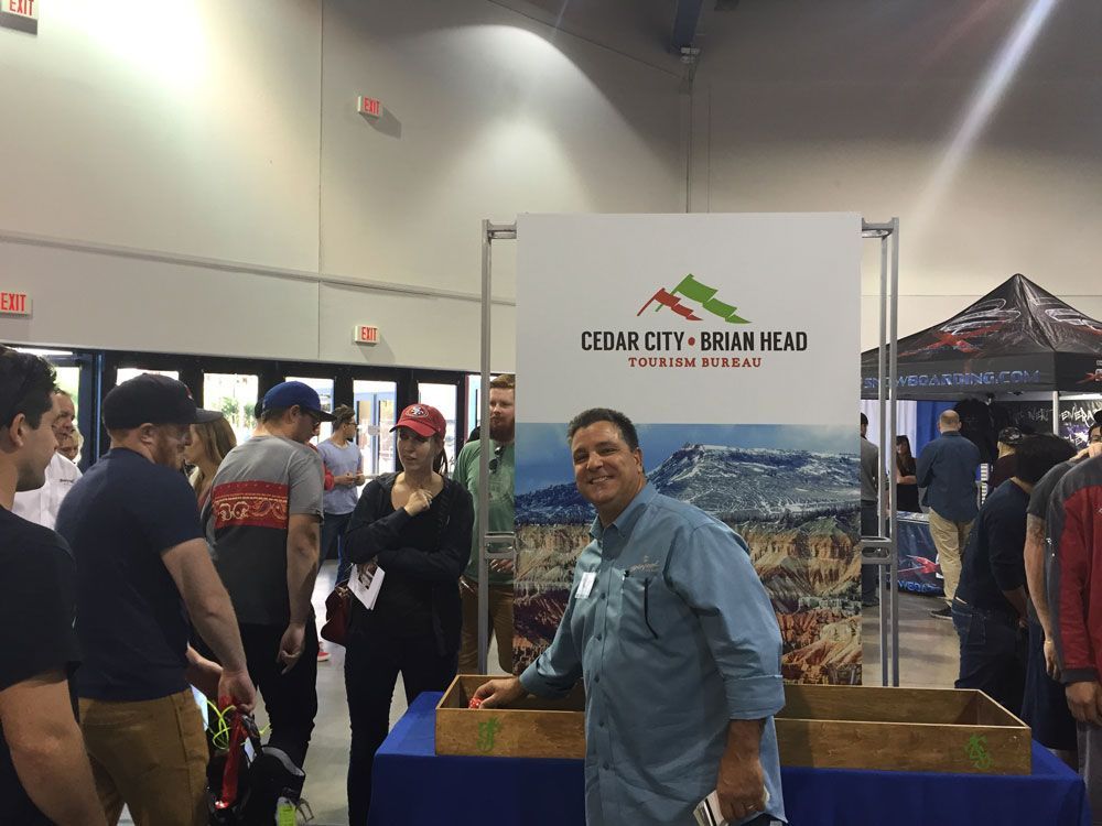 Man smiling at a Cedar City–Brian Head booth with a mountain display in a busy indoor expo