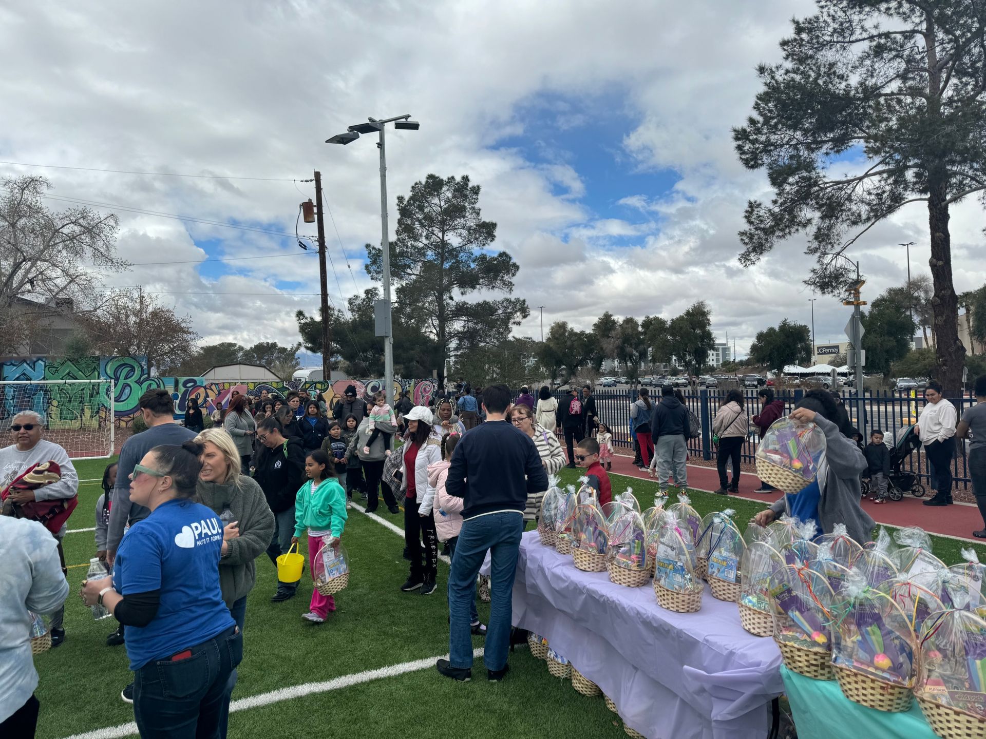 Crowd gathered in a park near tables covered with gift baskets under a cloudy sky.