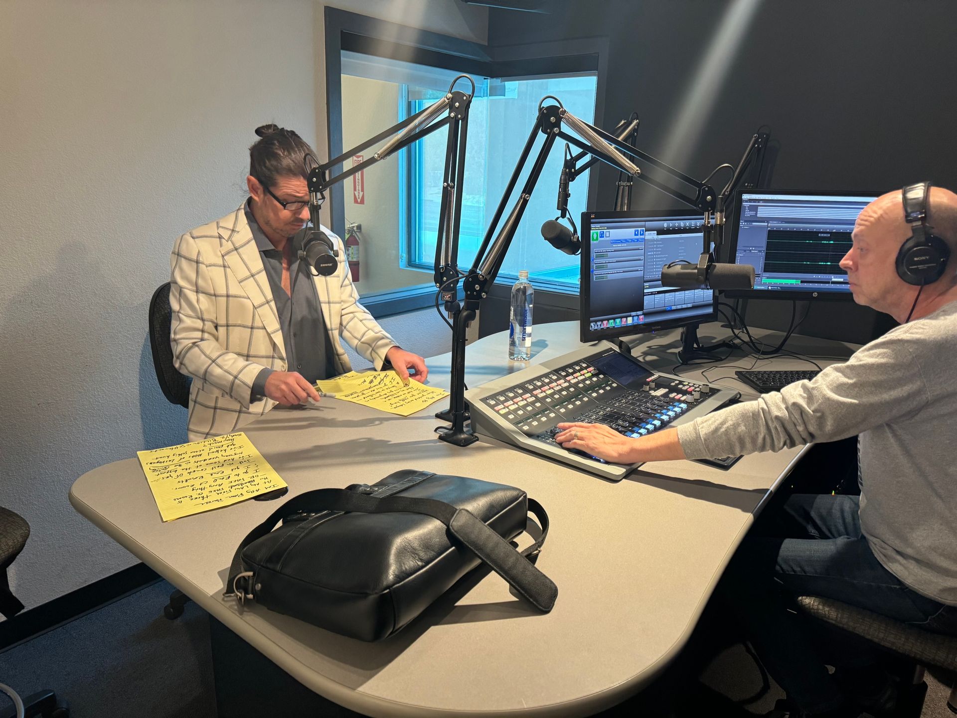 Two people recording a radio show in a studio, seated at microphones with audio equipment and monitors.