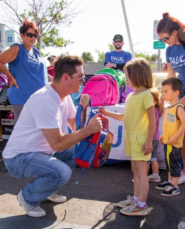 Man kneels to hand a blue item to two children at an outdoor event with volunteers and bags nearby
