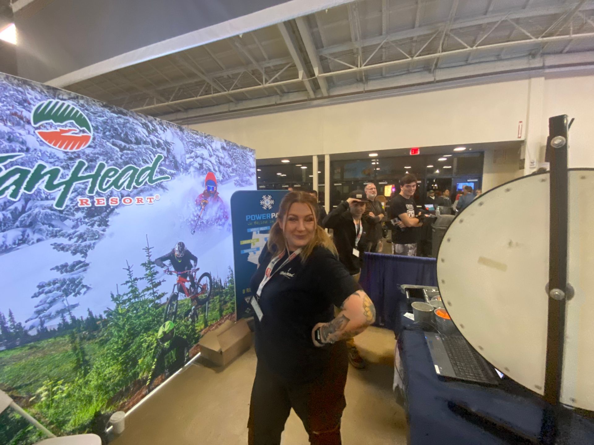 Woman posing at a trade show booth beside a large drum machine and mountain-themed backdrop.