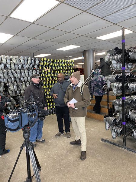 Two people talking in a sporting goods store aisle beside racks of shoes and gear, with a camera tripod in front.