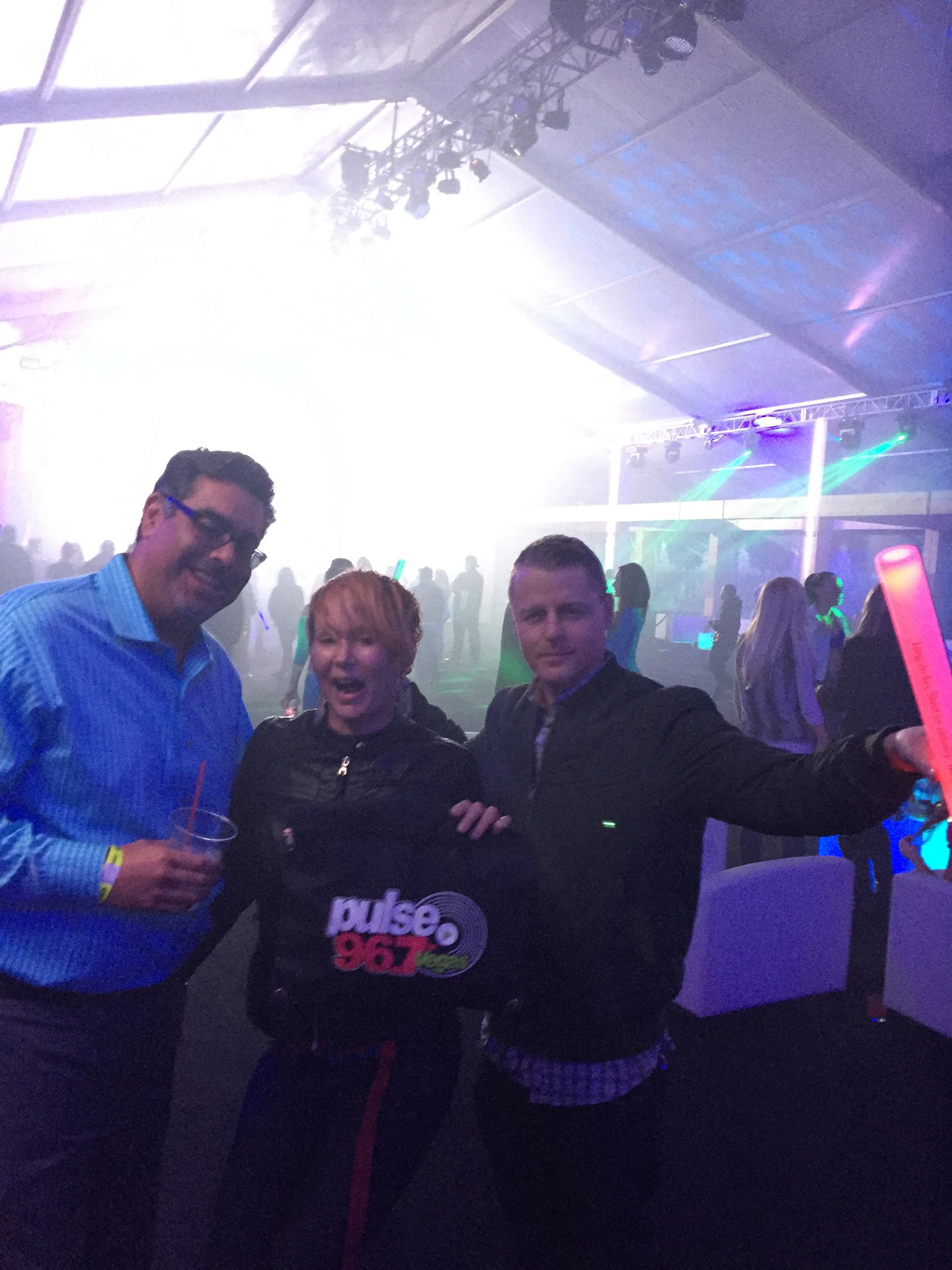 Three people posing and smiling in a brightly lit nightclub, with colorful lights and a bar in the background
