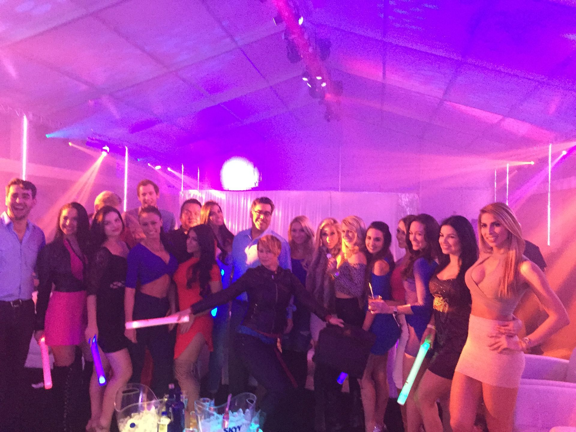 Group of people posing at a neon-lit party under purple lighting, some holding glow sticks.