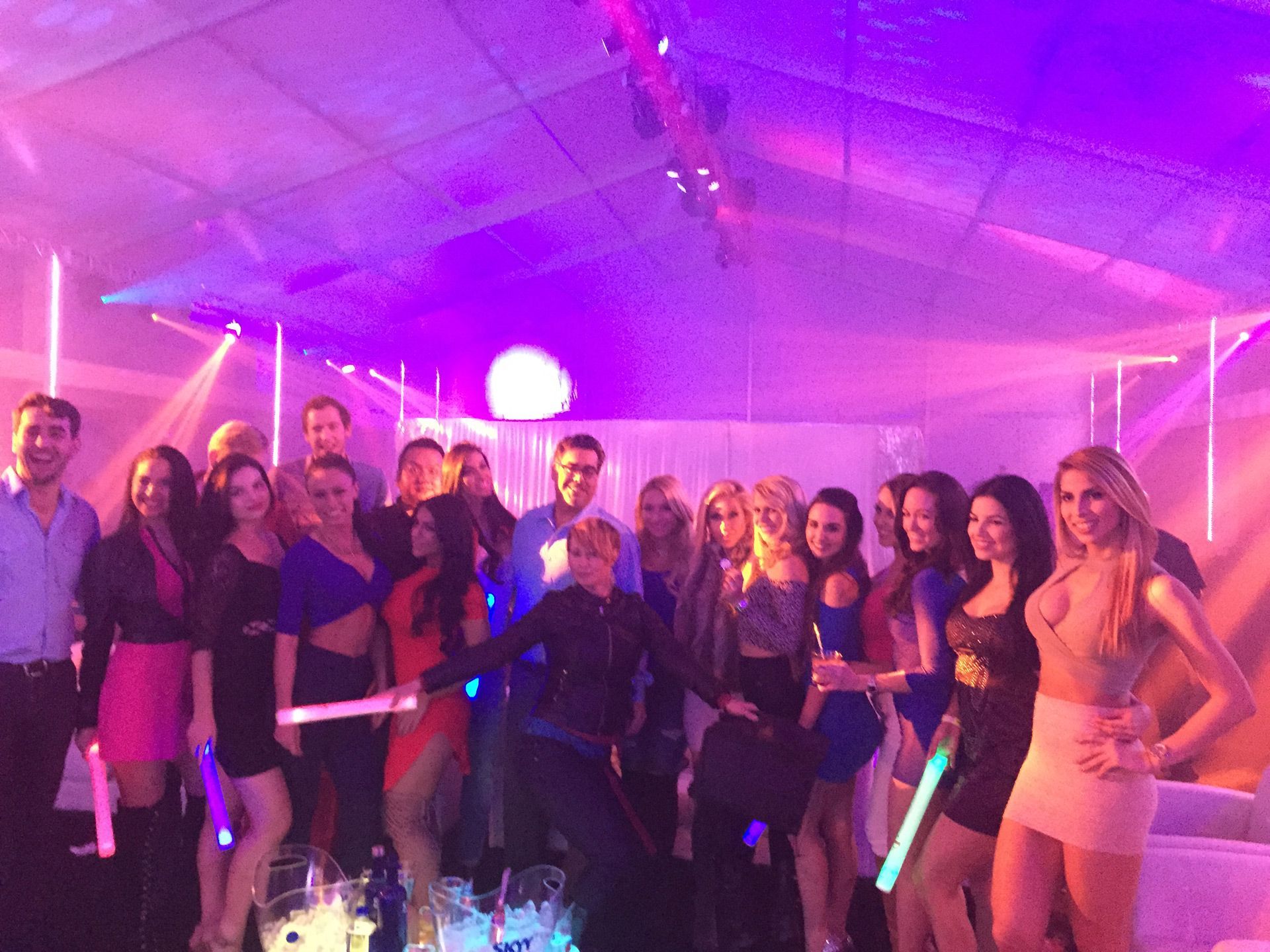 Group posing with glowing sticks at a purple-lit party inside a tent.