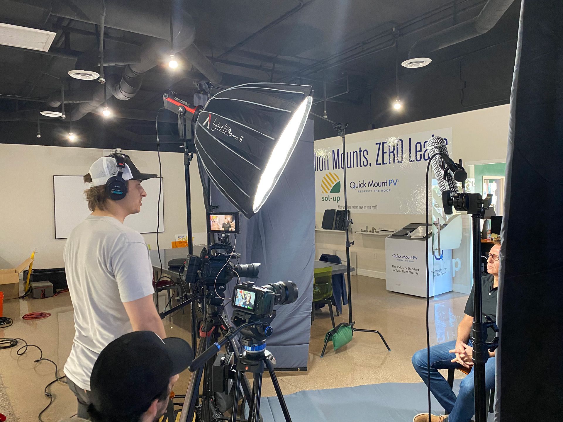 Film set with crew, camera, large softbox light, and seated subject in a studio-like space.
