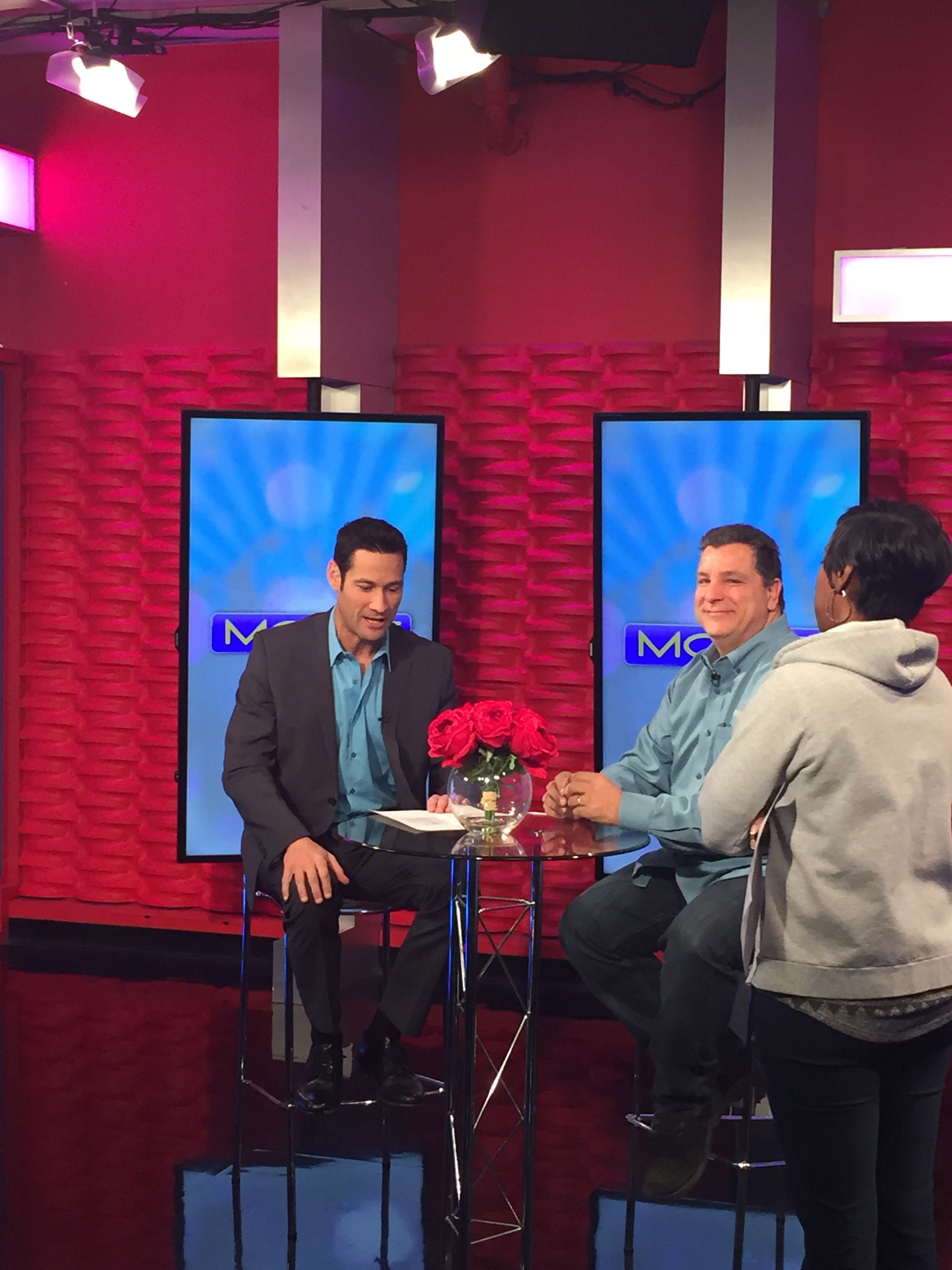 Two men sit at a studio table with flowers, as a person films them in a red-lit TV set.