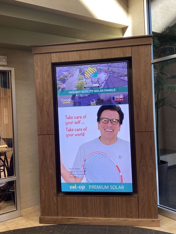 Digital ad display in a wood kiosk, showing a smiling man and Spanish text for “lentes prev/solares” campaign