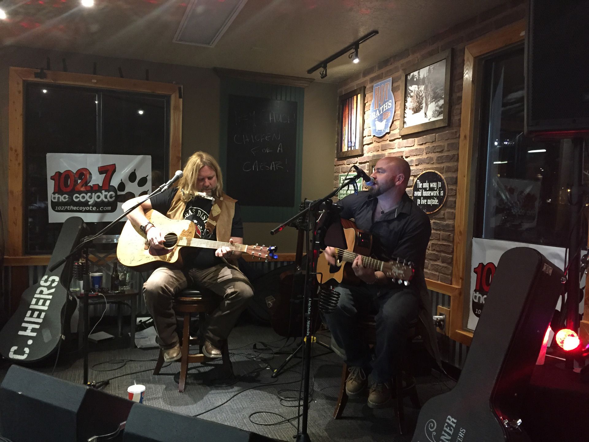 Two musicians playing acoustic guitars on a small indoor stage, with microphones and warm bar lighting.