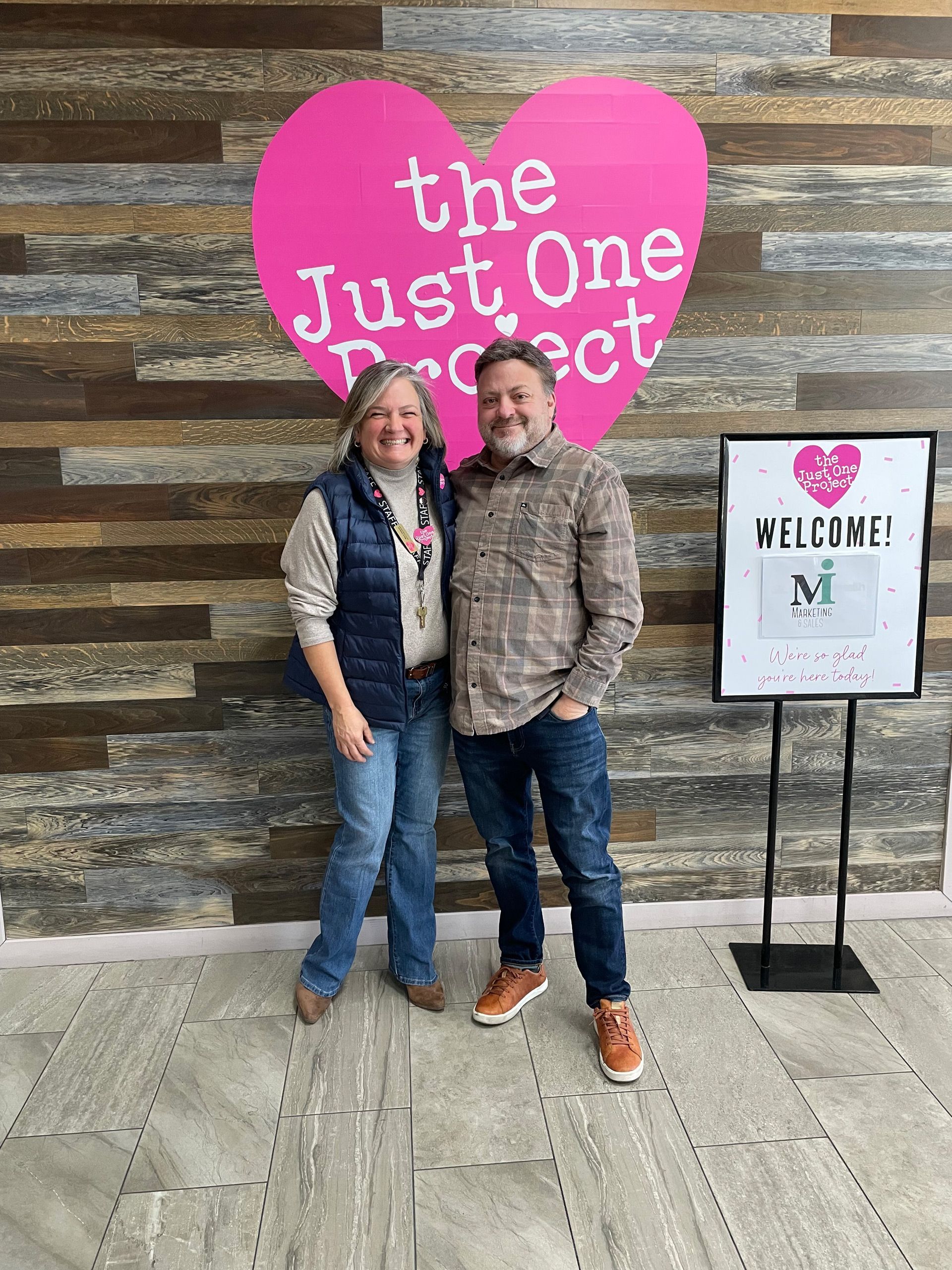 Two people pose by a pink “The Just One Project” sign and a “Welcome” stand in front of a wood wall.