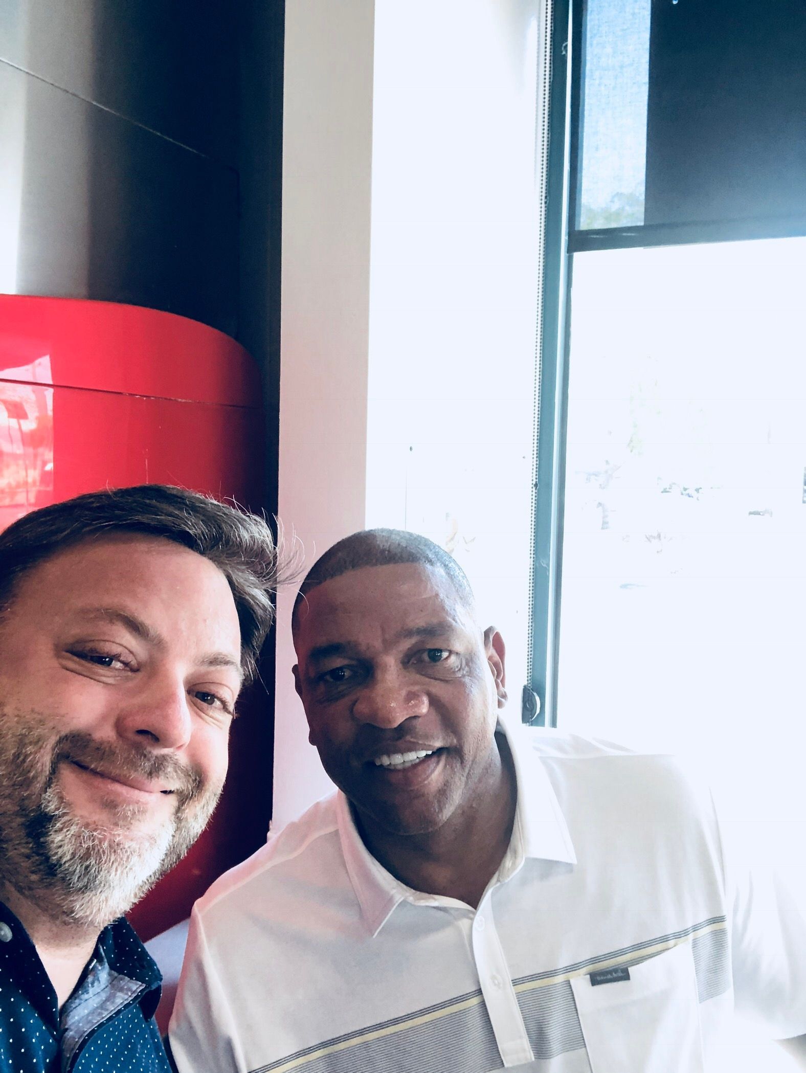 Two men smiling for a selfie indoors near a bright window