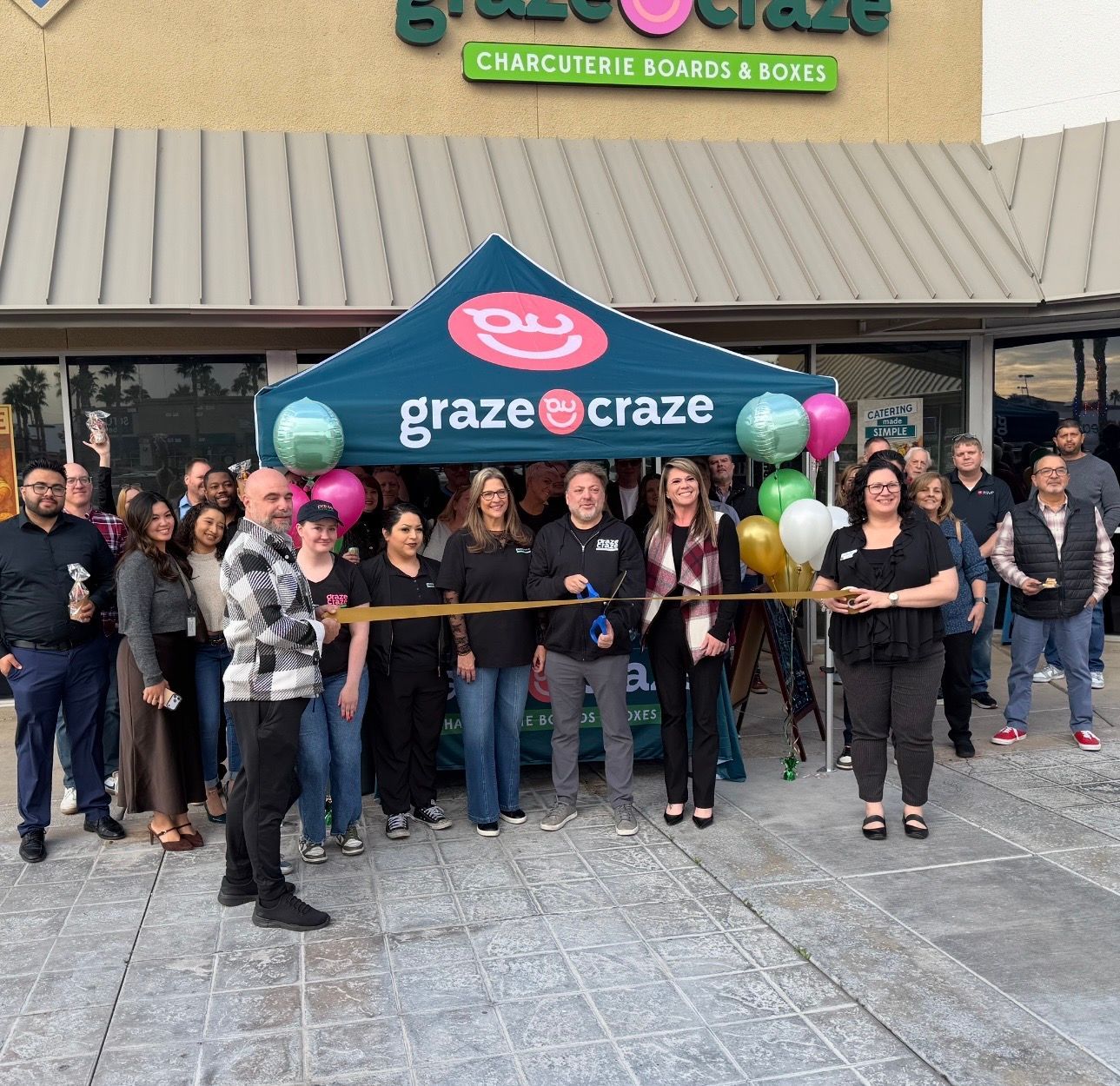 Group of people posing outside a Graze Craze storefront under a teal tent with balloons and a ribbon-cutting setup