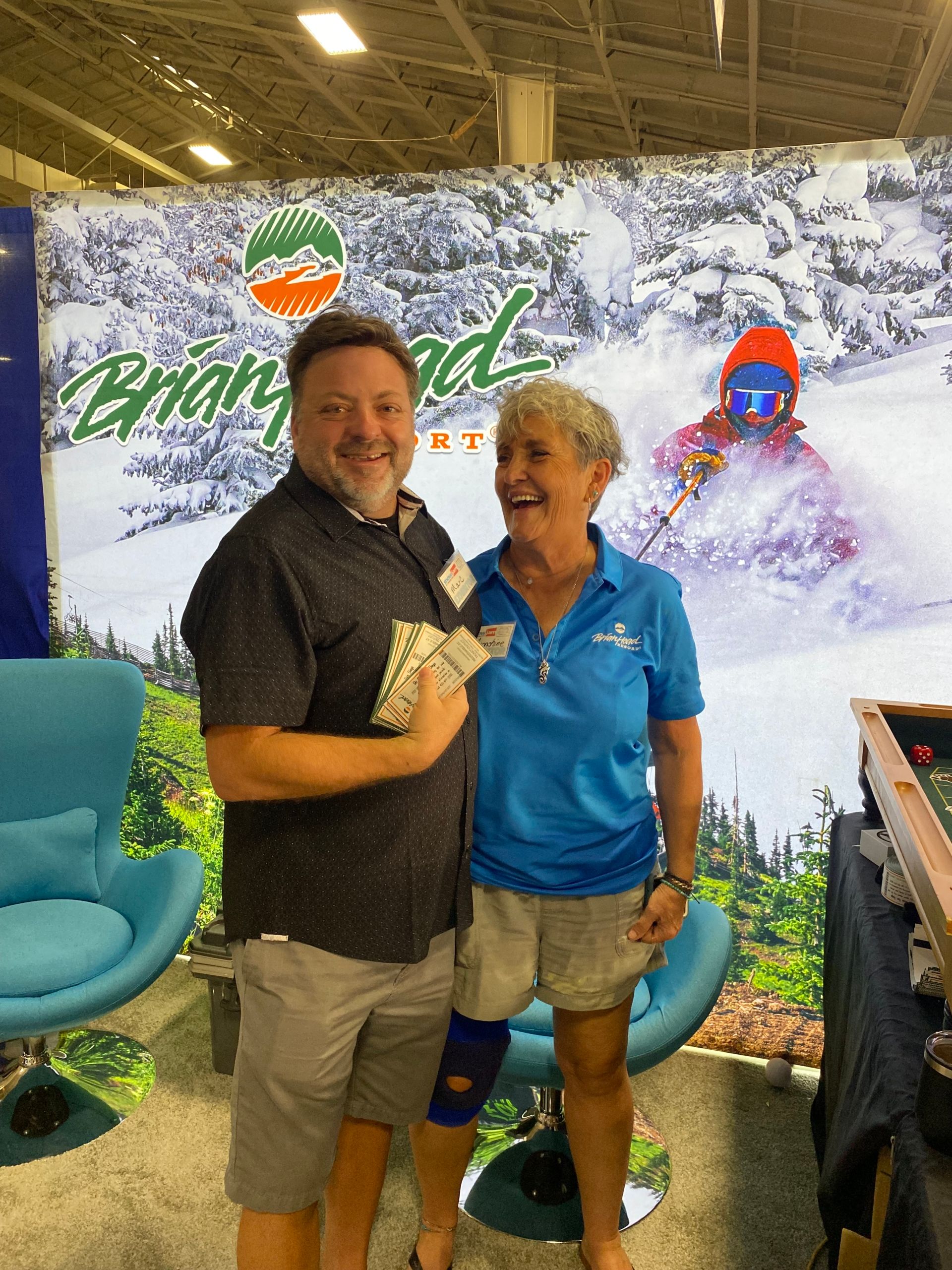 Two smiling people pose at a trade show booth with a green-and-white backdrop and blue chairs.