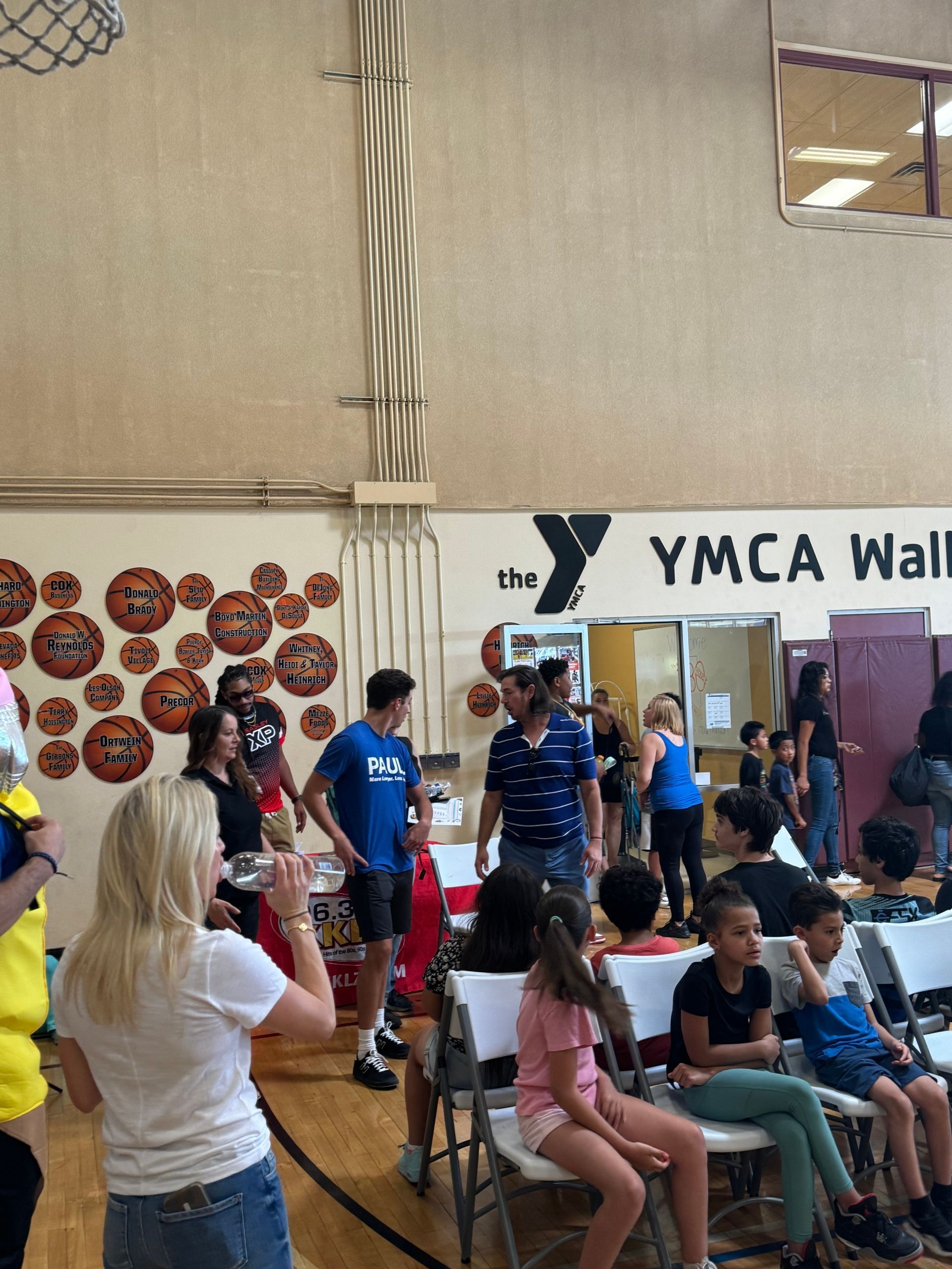 People gathered in a gym near a wall labeled “YMCA Wall,” with seated and standing spectators.
