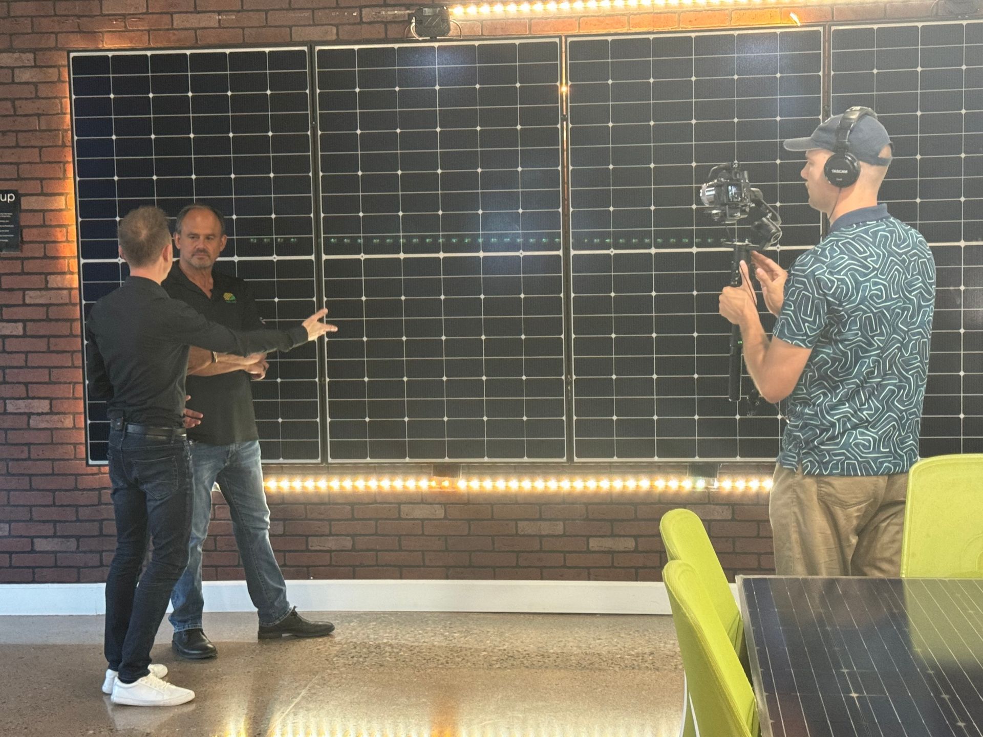 Two men and a cameraman in a brick-walled studio, with one man pointing at a large wall display.