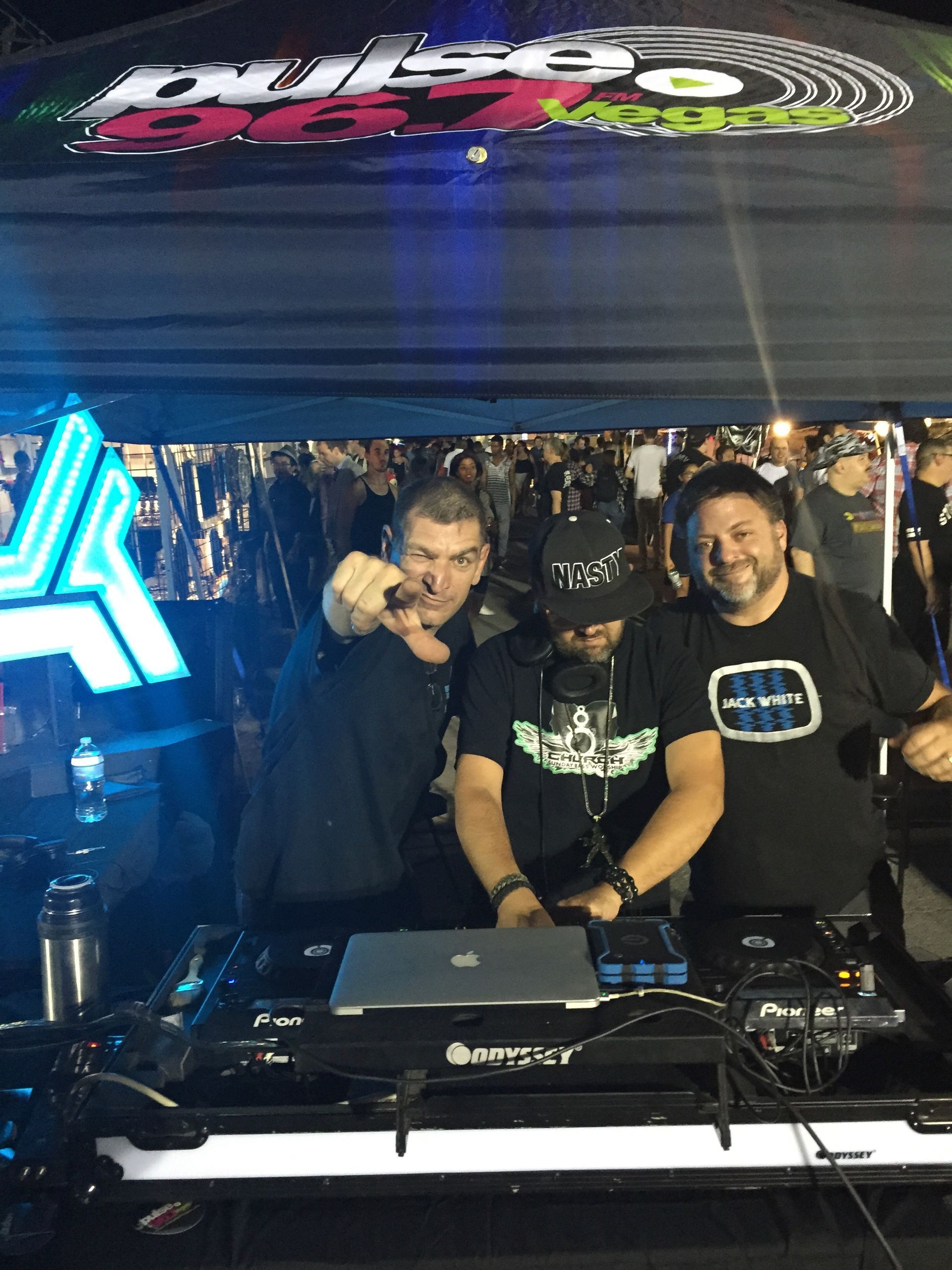 Three DJs smiling behind a mixer under a black tent with a neon sign above them.