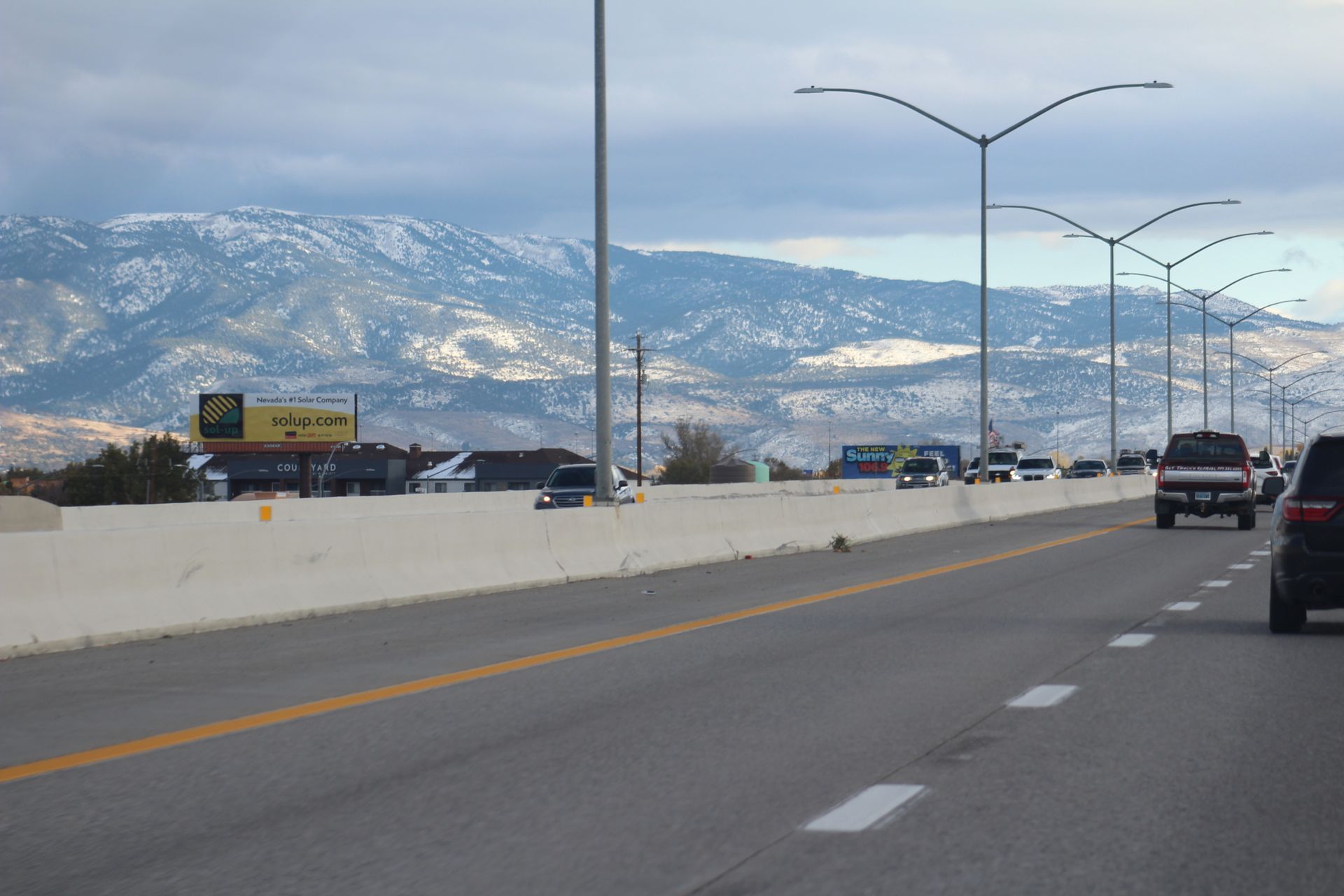 Highway with cars and a concrete barrier, mountains in the snowy distance under a cloudy sky