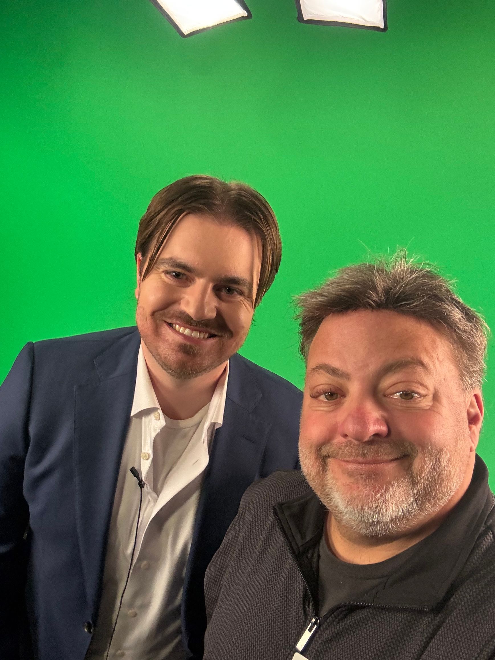 Two men smiling in front of a green screen, one in a blue blazer and one in a black jacket.