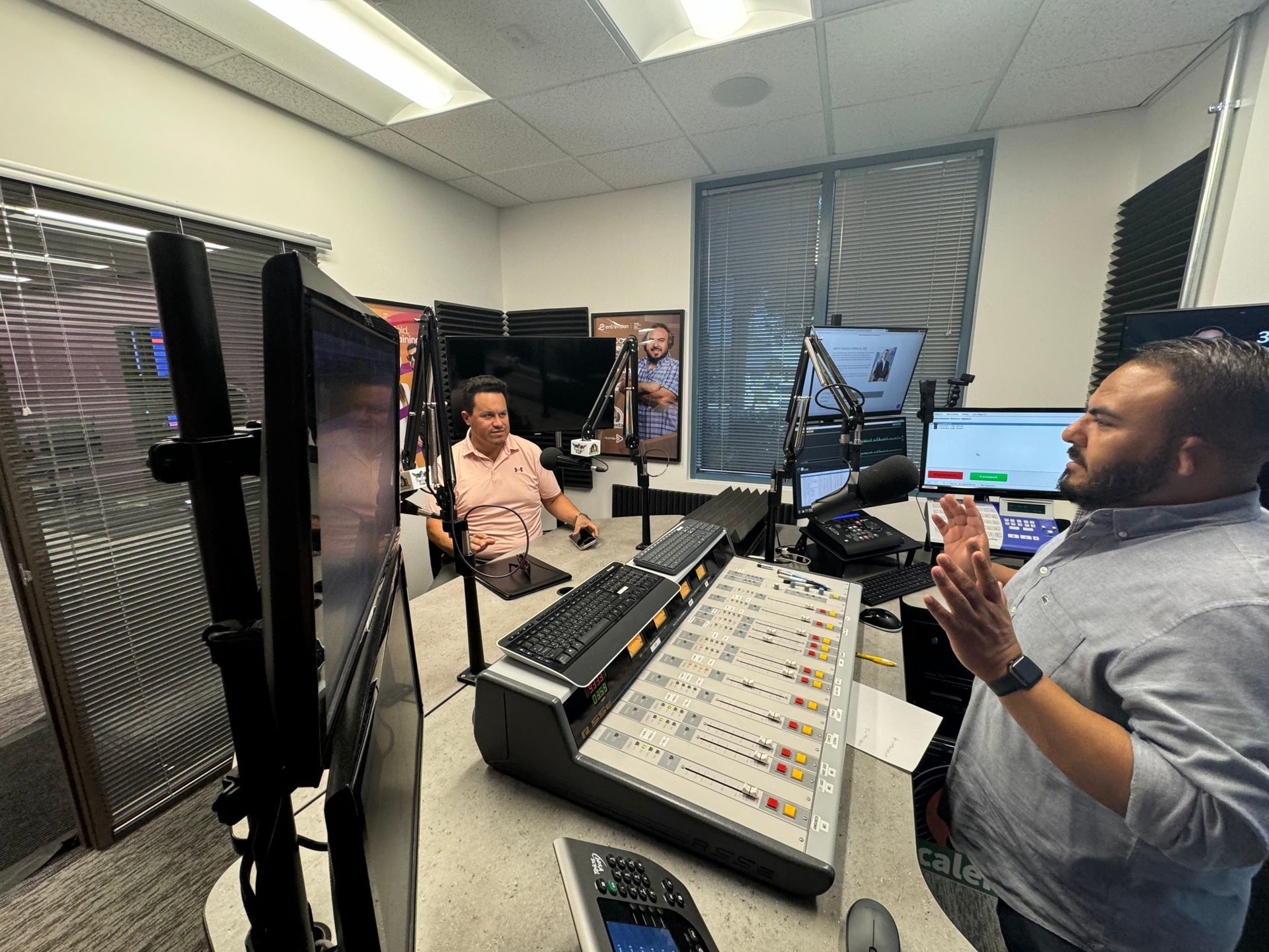 Two people in a radio studio with microphones, mixing console, and computer monitors