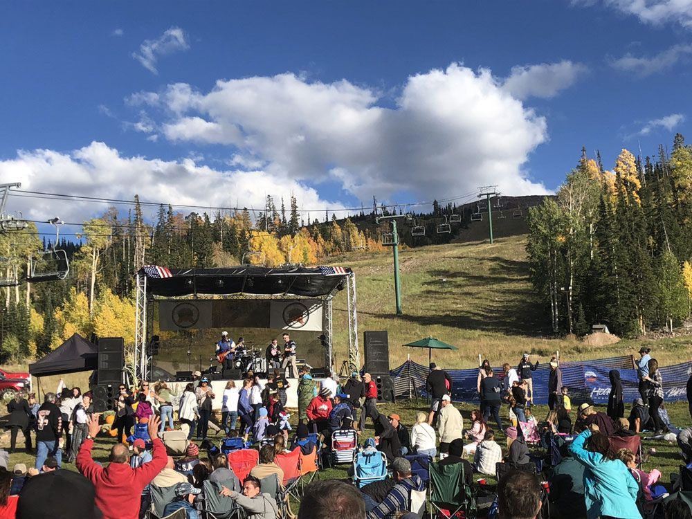 Crowd watching an outdoor concert on a mountain stage under a blue sky