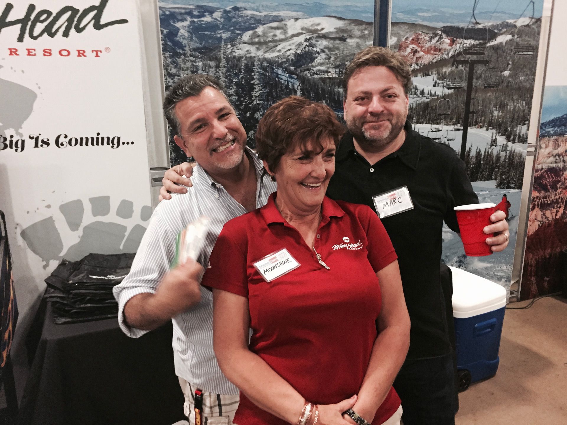 Three people smiling at a Head Resort booth, with a snowy mountain backdrop and red drinks in hand