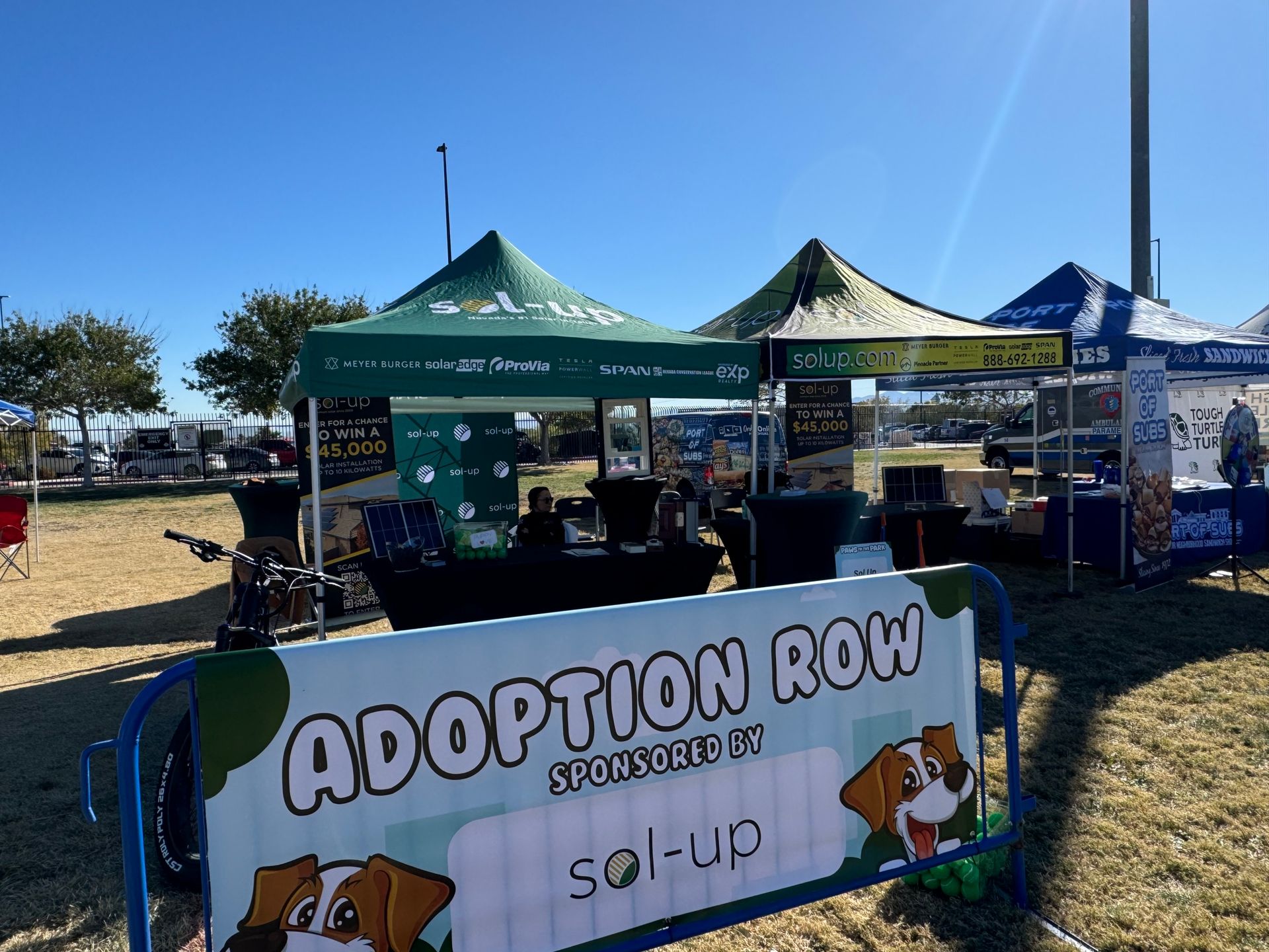 Adoption row booth at an outdoor event with green tents and a white sign, under a bright blue sky