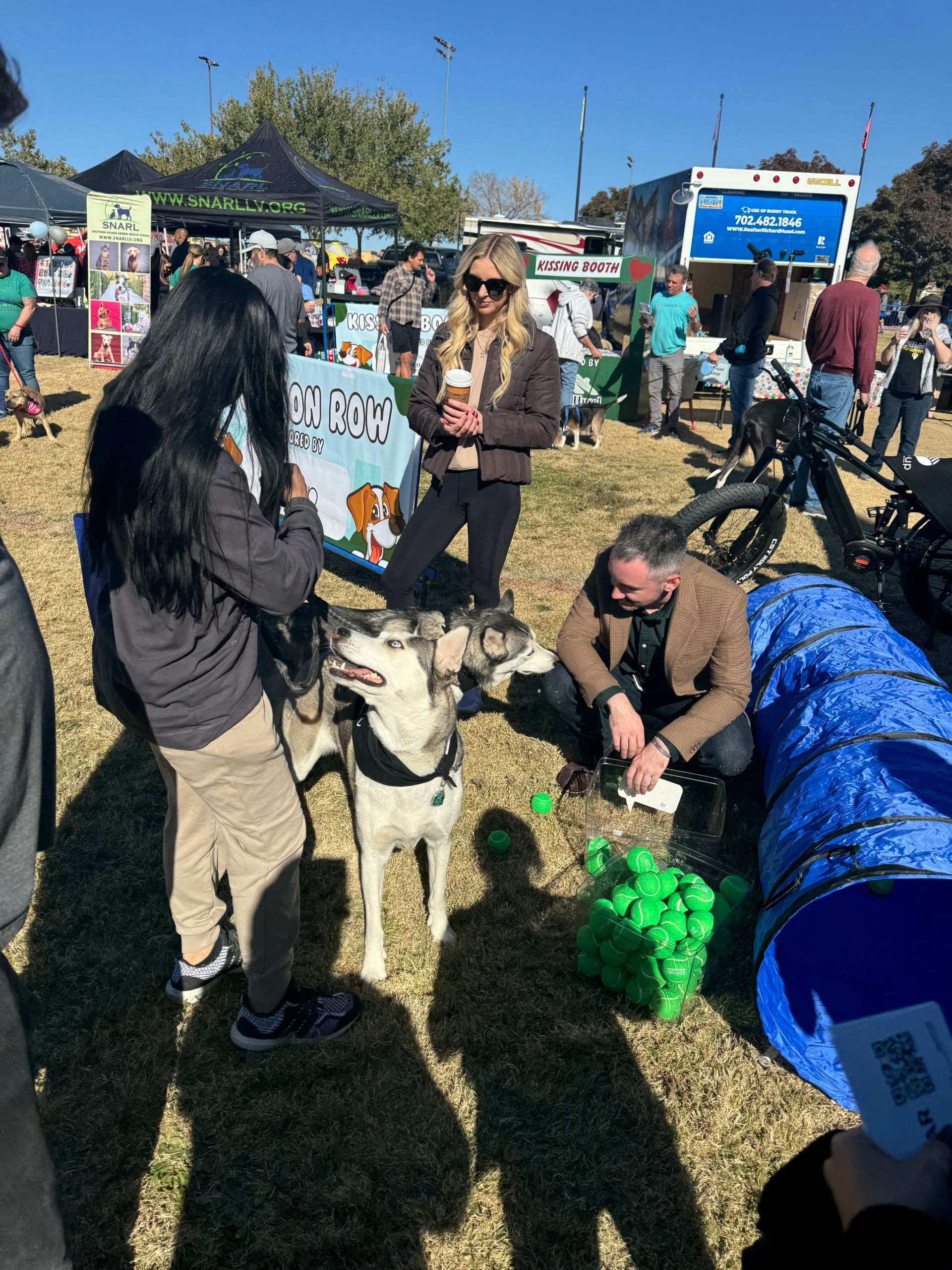 People gathered at an outdoor fair, petting a husky beside a booth on a sunny day.