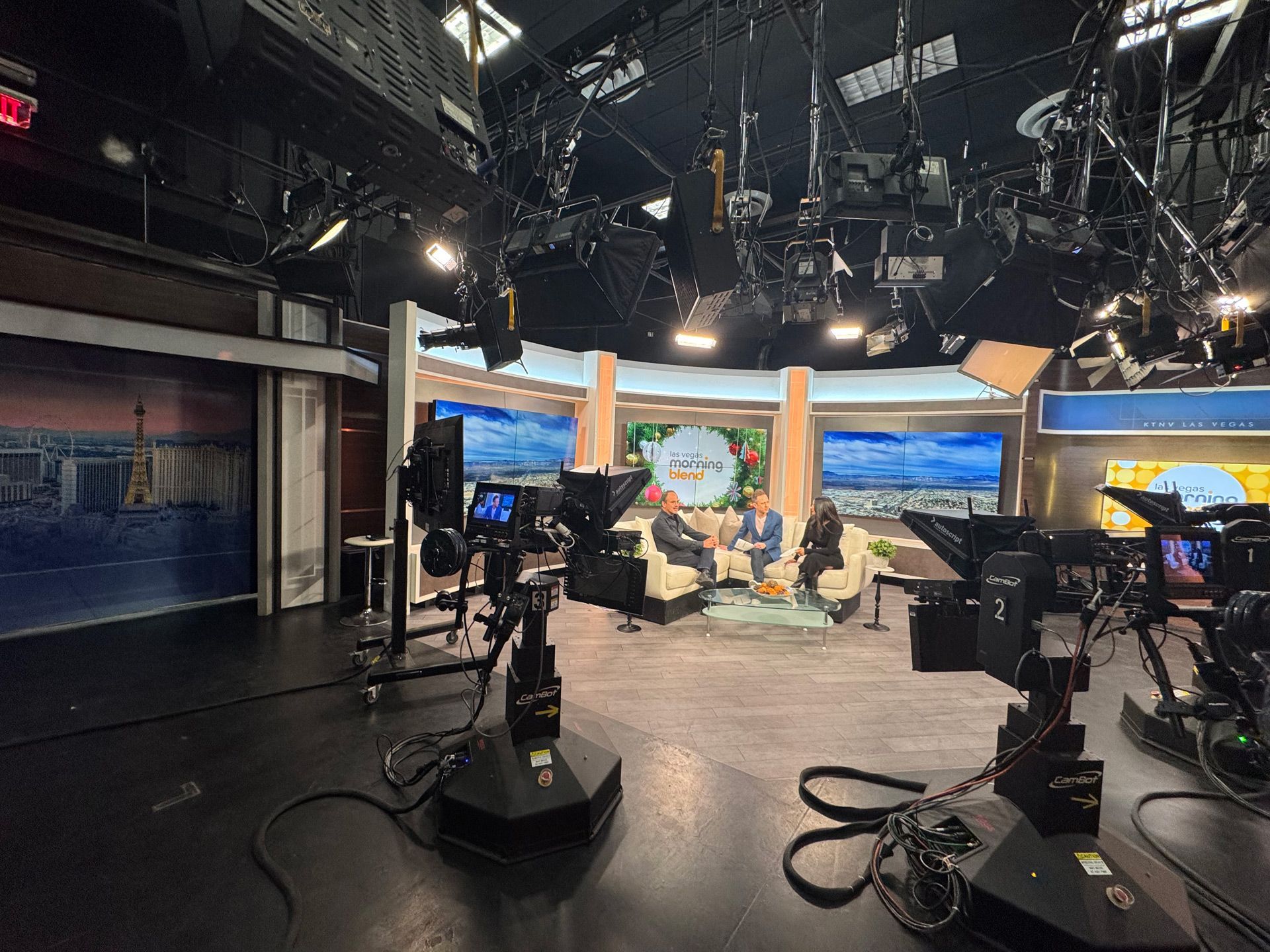 TV studio set with cameras and lighting, showing a cityscape backdrop and seated hosts at center stage