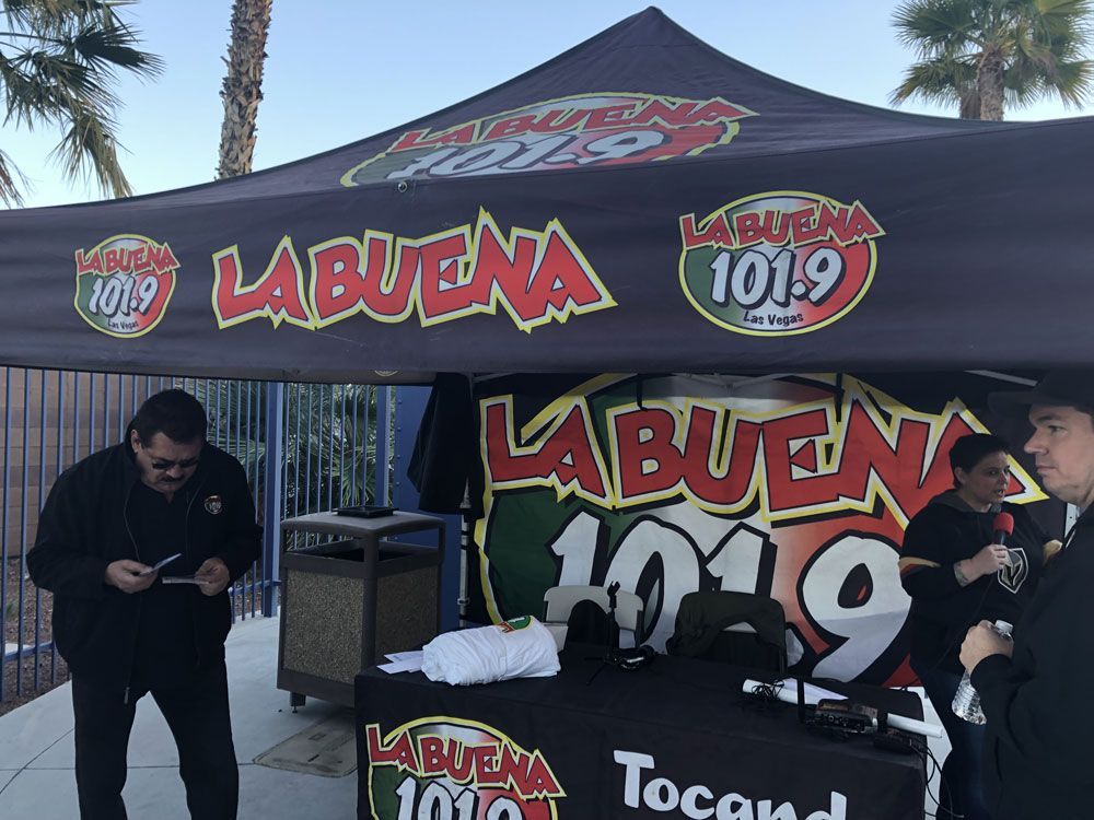 LA BUENA 101.9 radio booth tent at an outdoor event, with two people standing nearby
