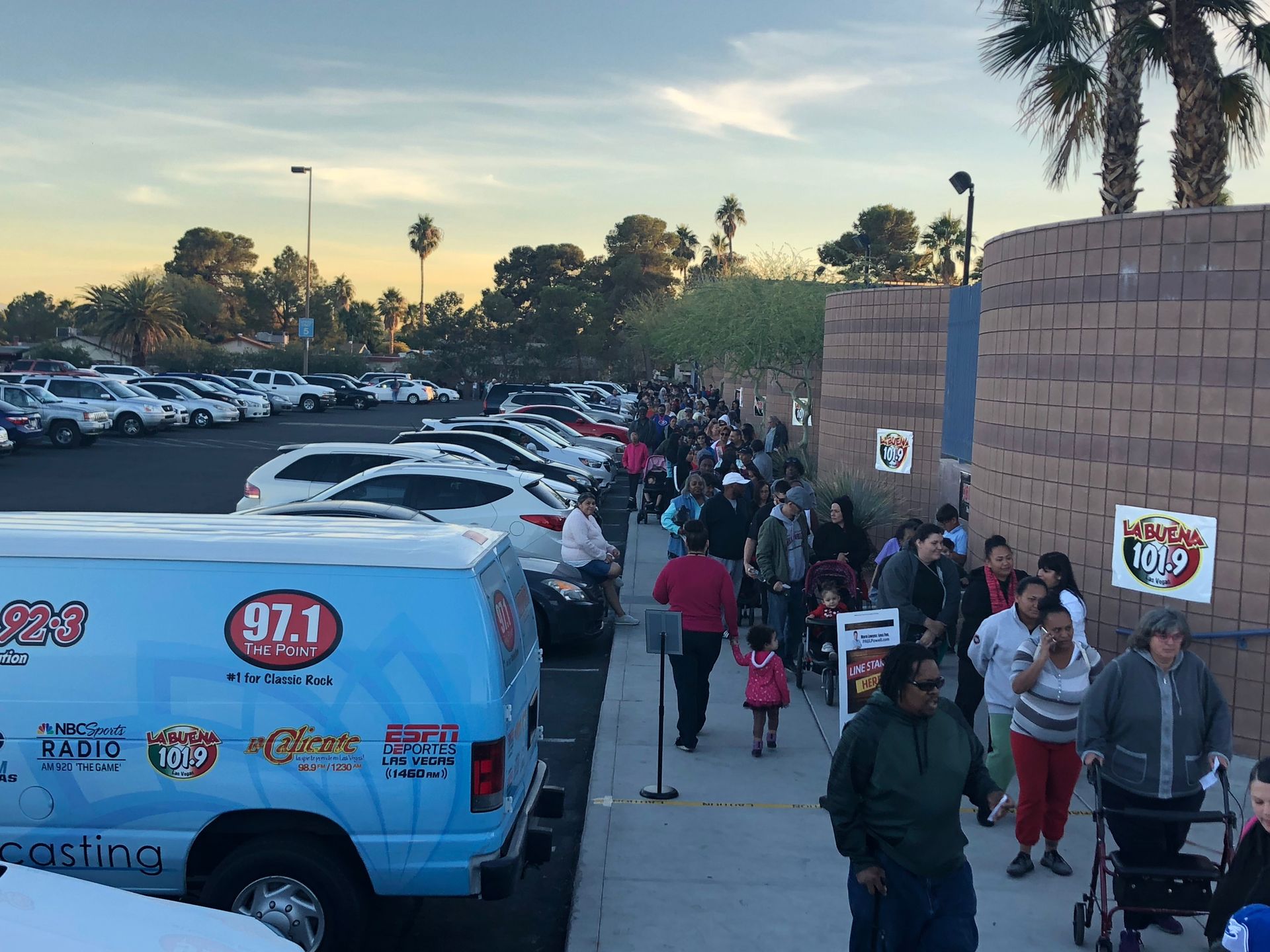 People lined up outside a building beside a blue van and parked cars at dusk