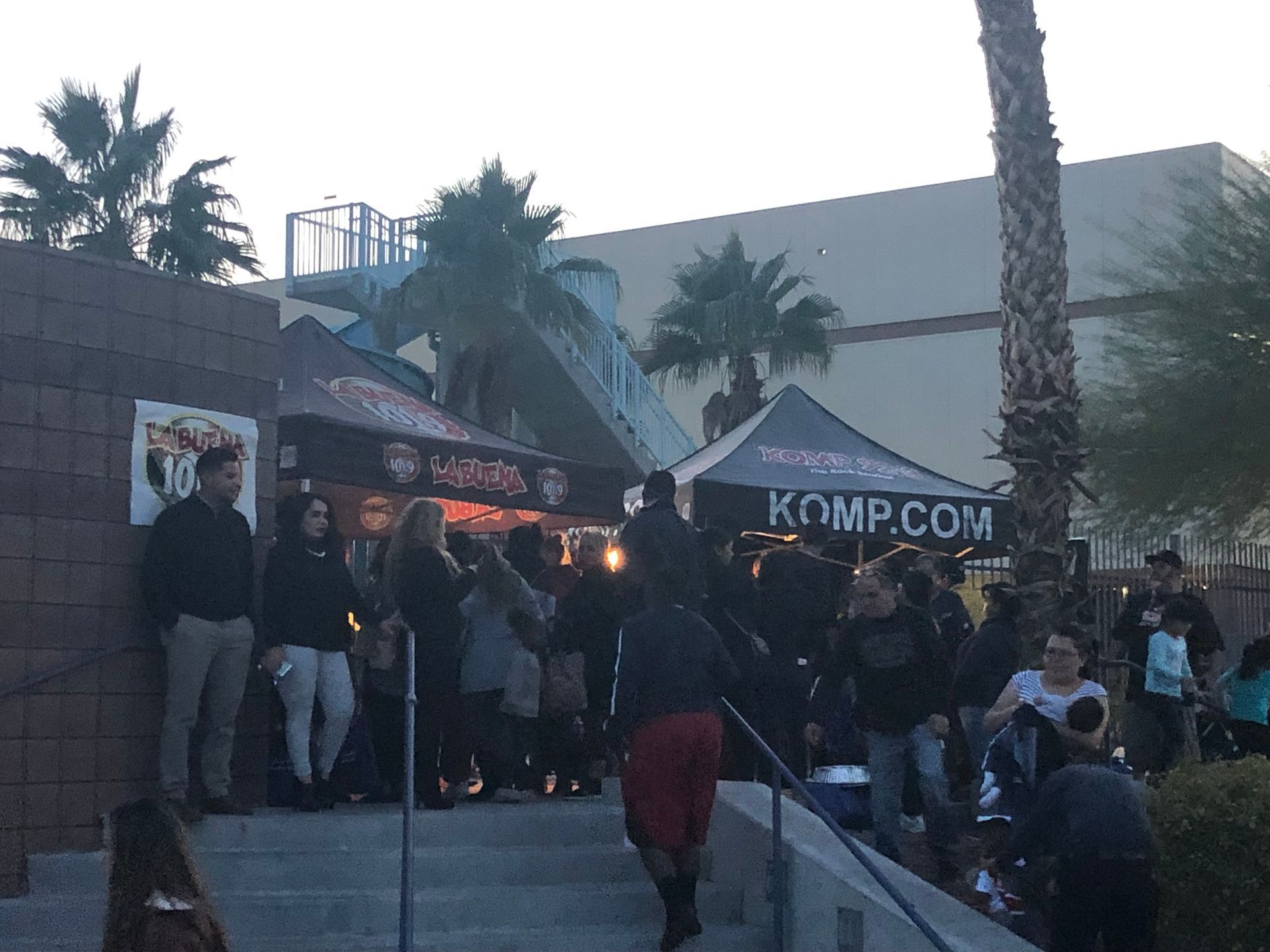 People gathered outside a building near tents and palm trees at dusk.