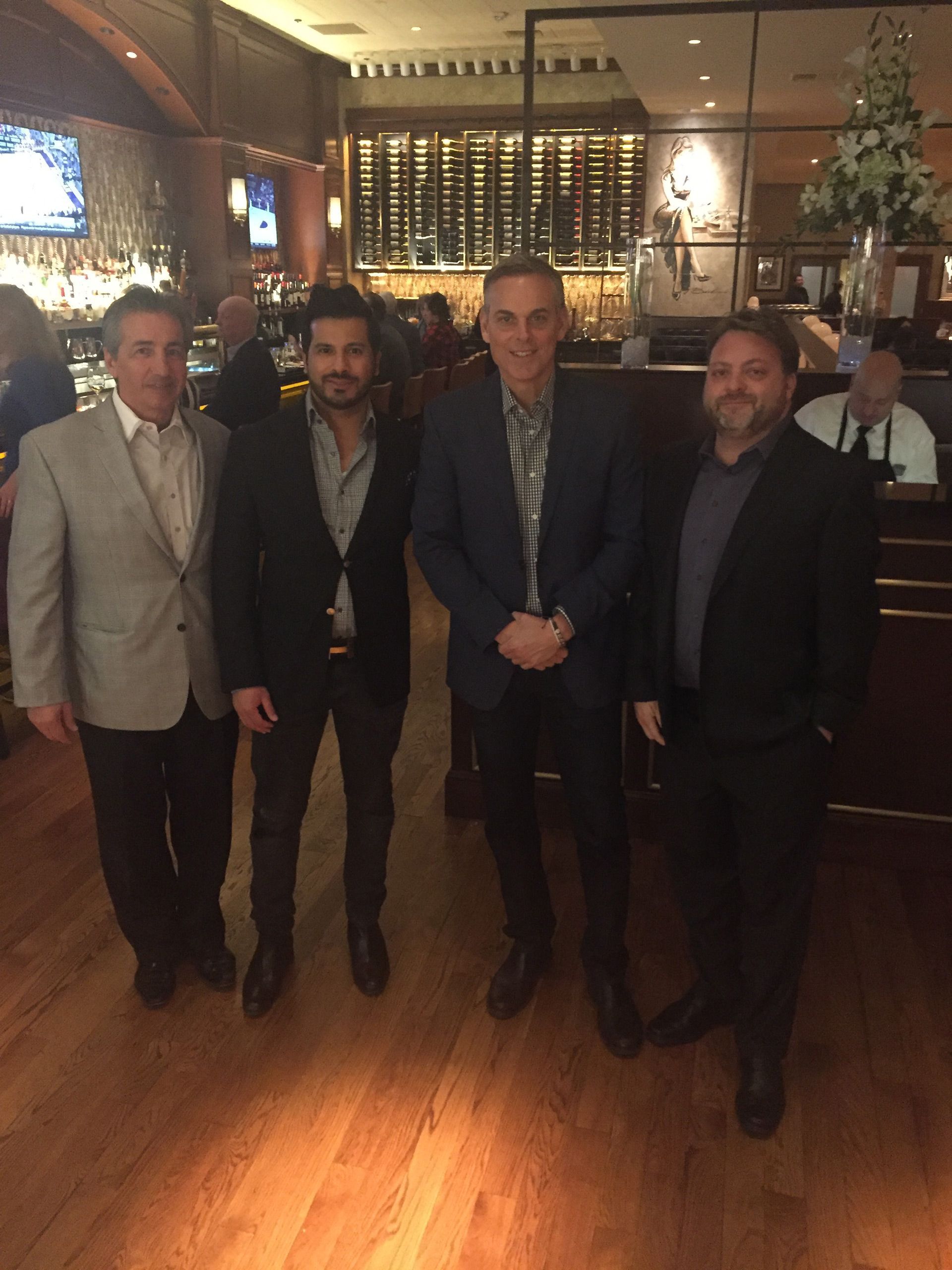 Four men posing together in a warmly lit bar or restaurant with wooden floors and shelves behind them