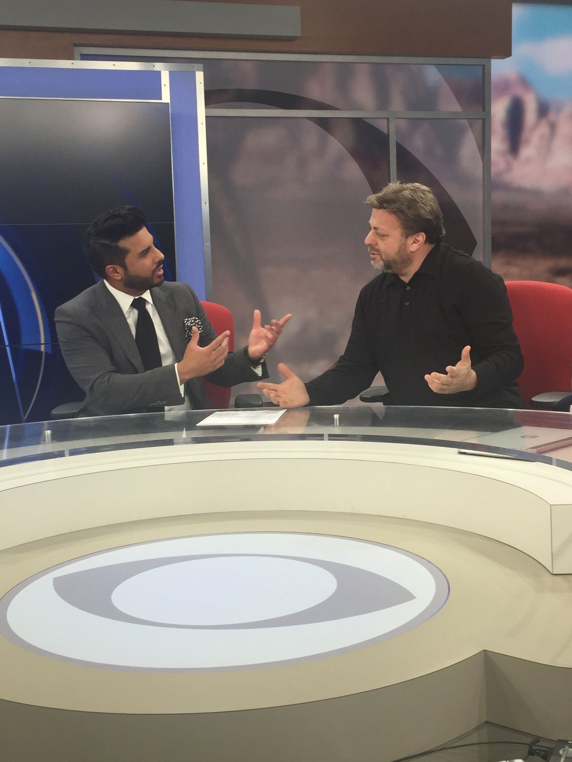 Two men in conversation at a news desk, with a blue and white studio backdrop.