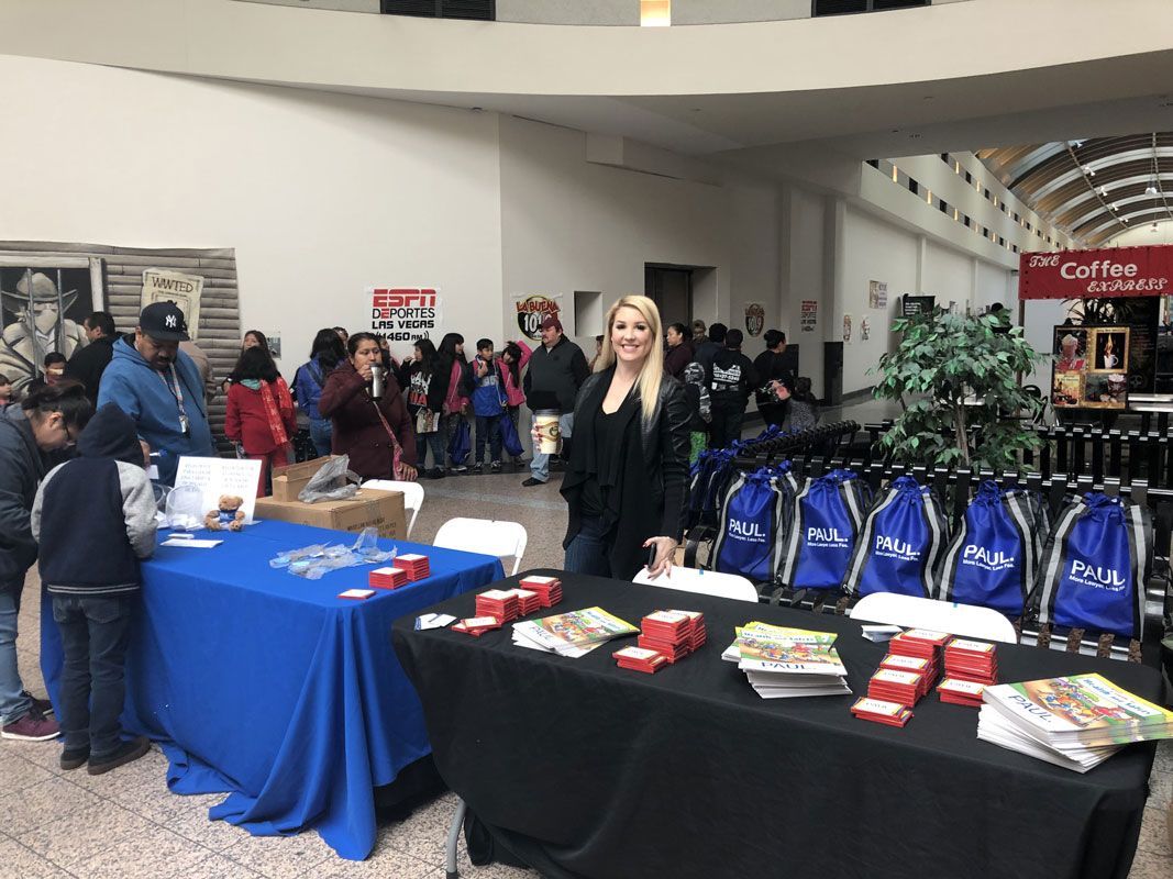 Trade show booth with tables of red-and-white items and blue bags, with a woman standing behind the display.