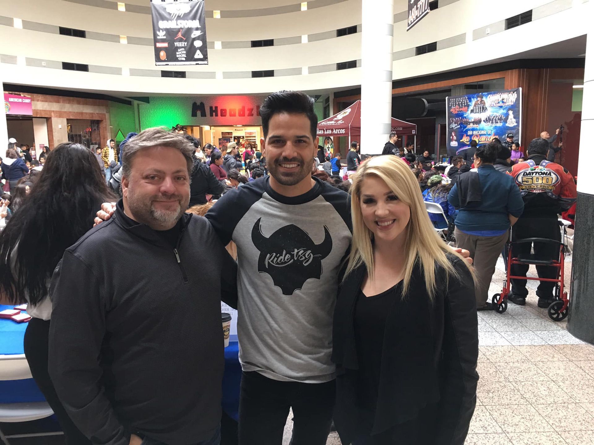Three people smiling indoors at a busy shopping mall or event venue