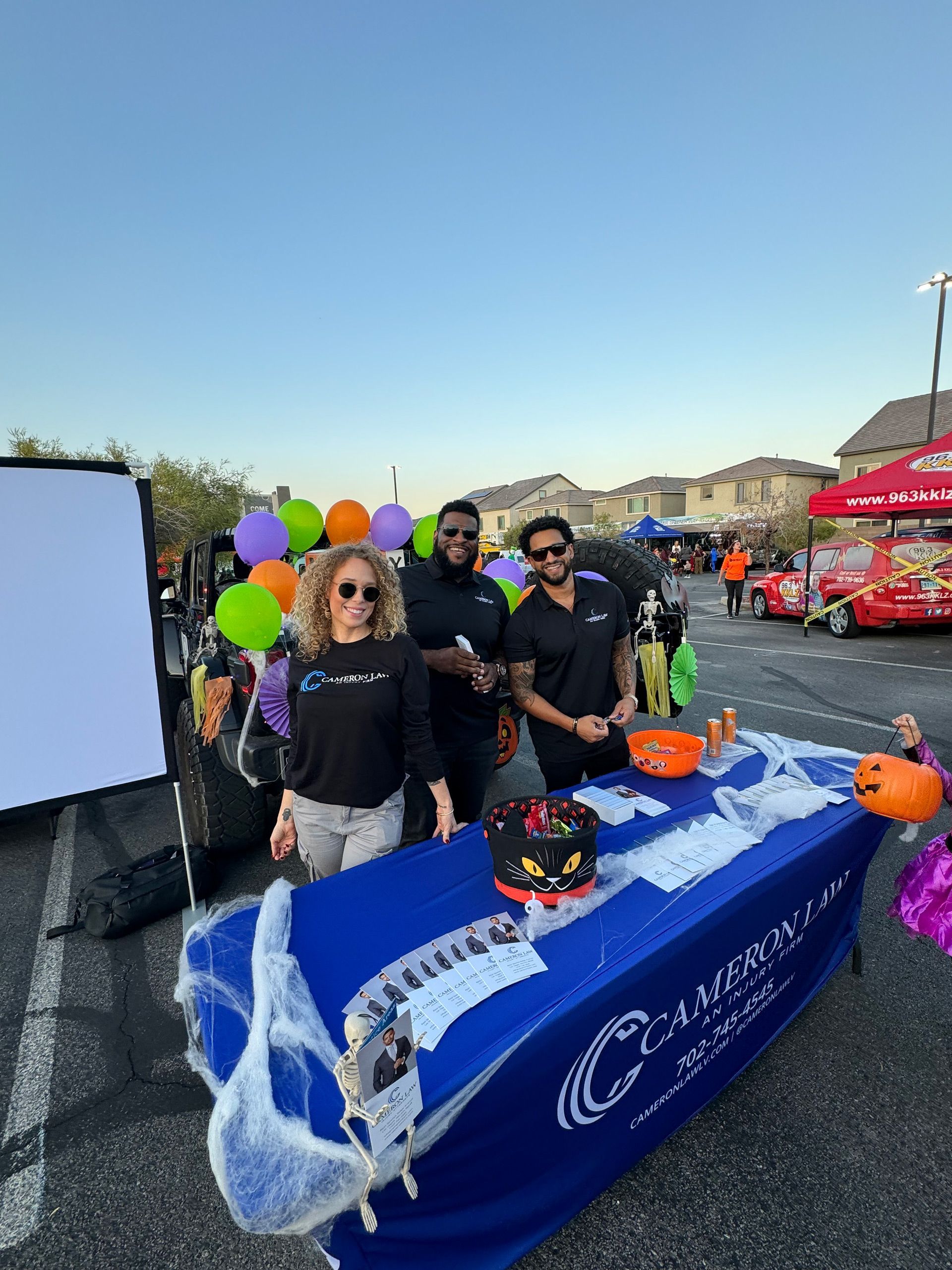 People behind a blue parade table with balloons, Halloween decorations, and event tents along a street.