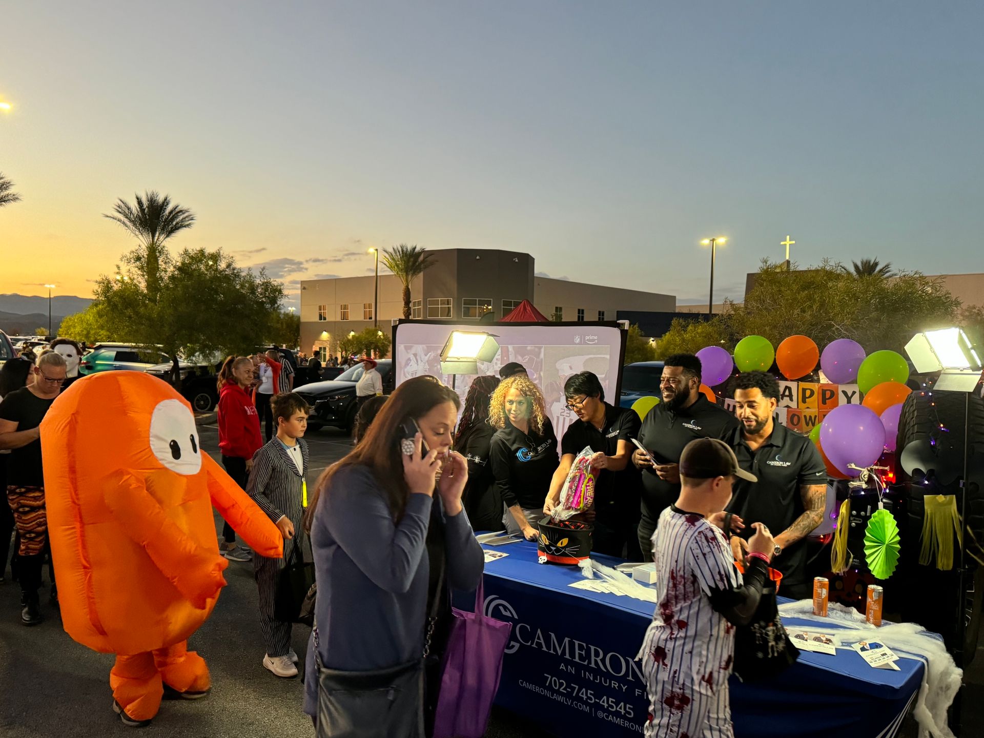 Outdoor festival booth with orange mascot, people gathering, and colorful balloons at dusk