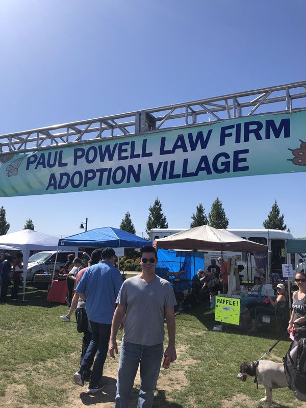 Outdoor event banner reading “PAUL POWELL LAW FIRM ADOPTION VILLAGE” above tents and people with dogs