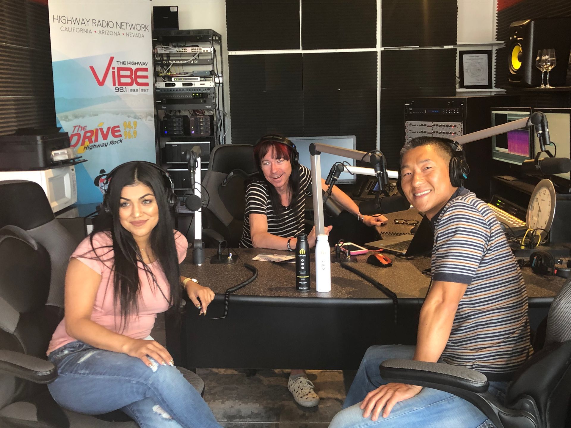 Three people seated at a radio studio table with microphones and headphones, smiling at the camera.