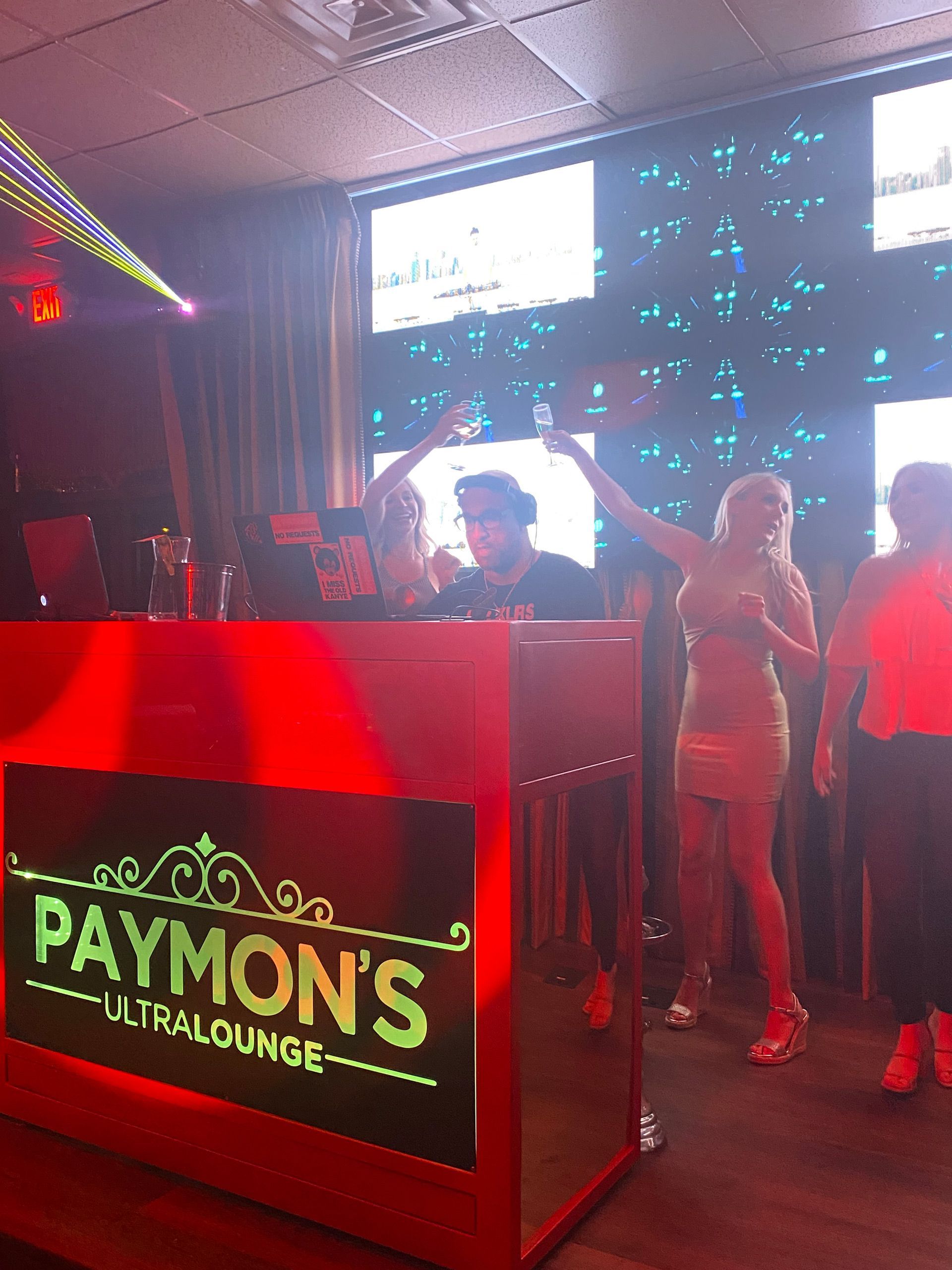 Nightclub DJ booth with neon “Paymon’s” sign and people dancing under red and blue lights