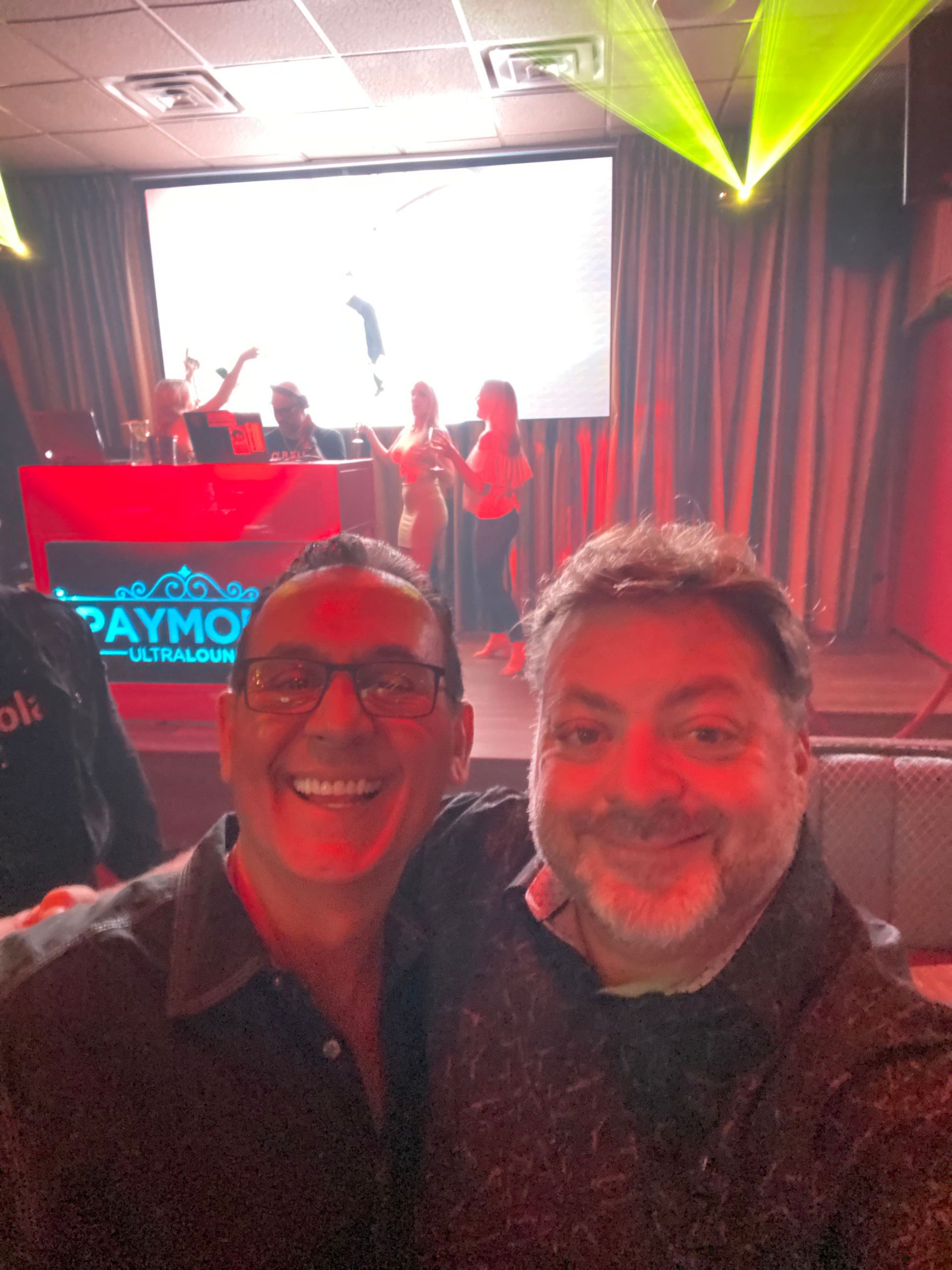 Two smiling people taking a selfie at a red-lit concert or nightclub stage