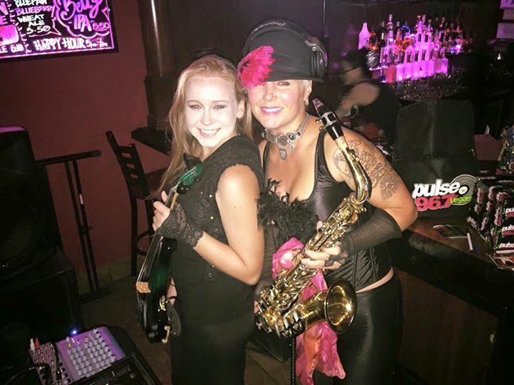 Two people posing in a dim nightclub, one holding a saxophone with pink accents.