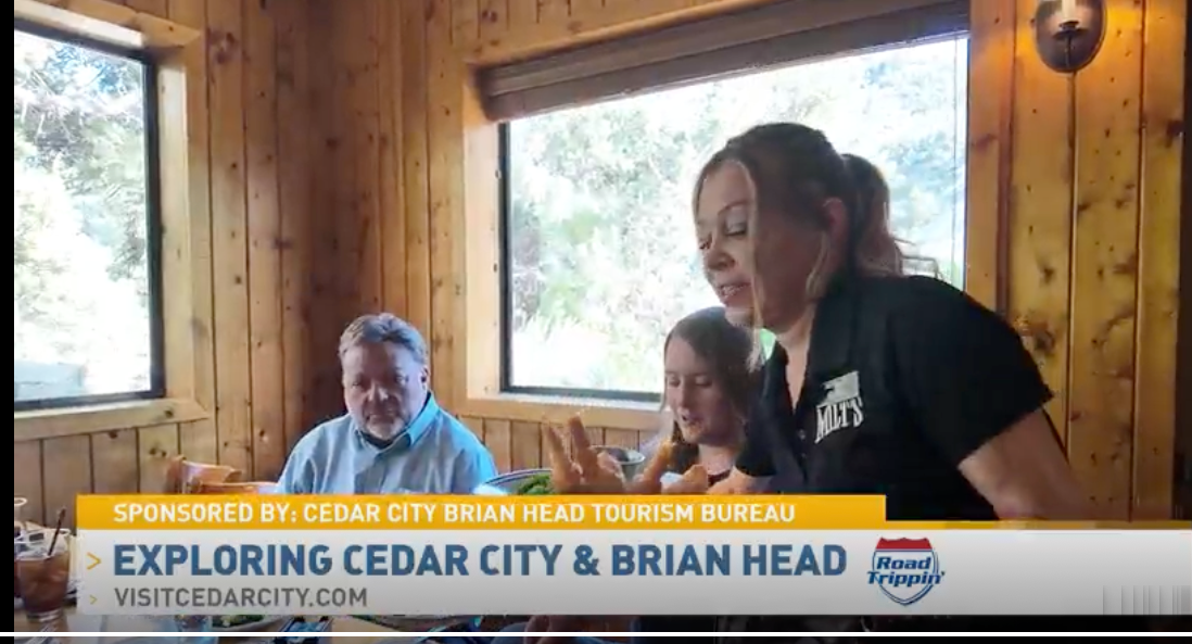 Three people in a wood-paneled room, smiling near windows, with a banner reading “Exploring Cedar City & Brian Head”