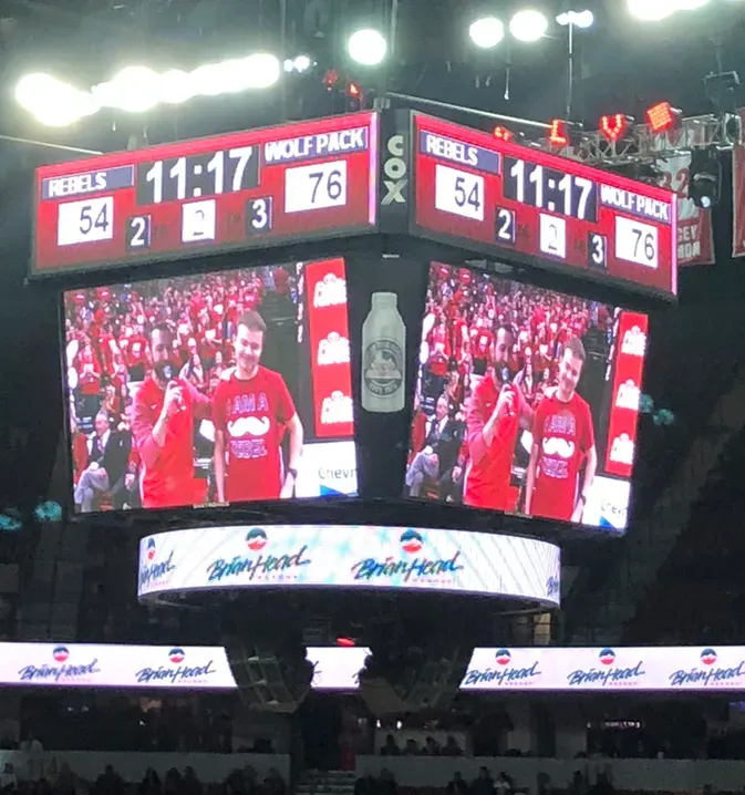 Basketball arena scoreboard shows Rebels leading Wildcats 76-54, with players on the jumbotron below.