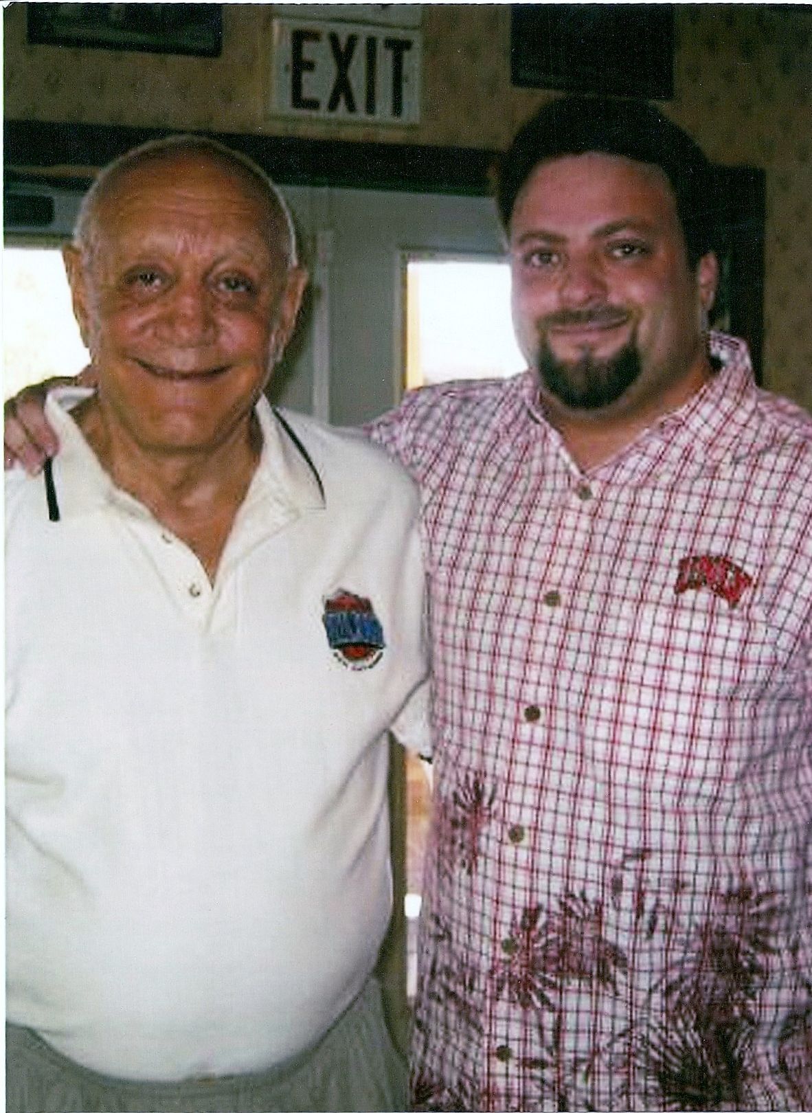 Two men standing together indoors, smiling, with an EXIT sign behind them.