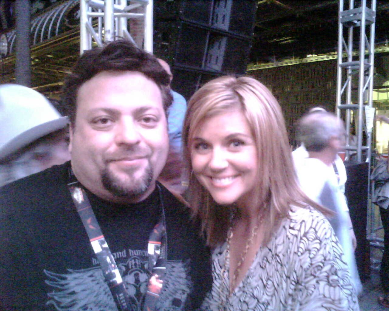 Two smiling people posing indoors at an event, with a stage and lights in the background.