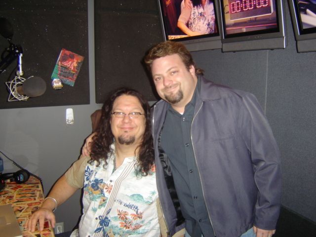 Two people smiling together in a dim room with framed posters on the wall.