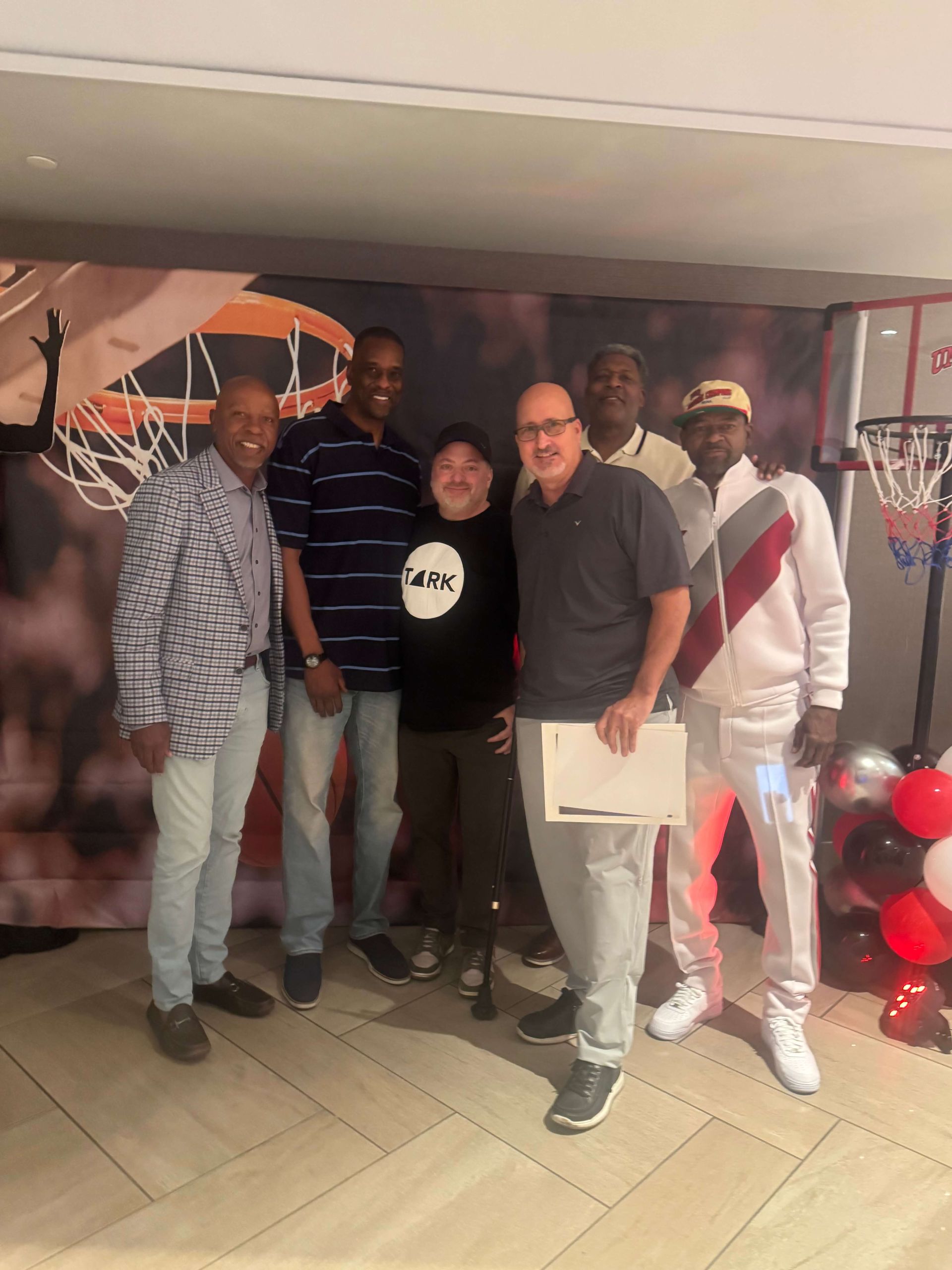 Six people posing indoors at a celebration, with balloons and a decorated wall backdrop.
