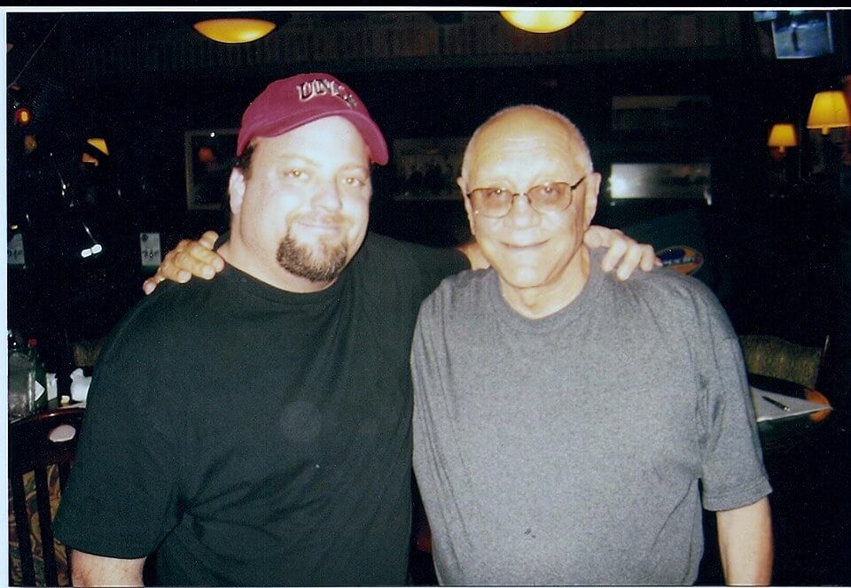 Two men posing together indoors, one in a red cap and black shirt, the other in glasses and a gray shirt.