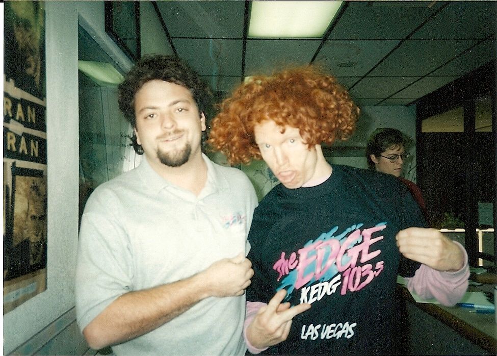 Two people posing in a hallway, one holding up a black T-shirt with pink and blue text.